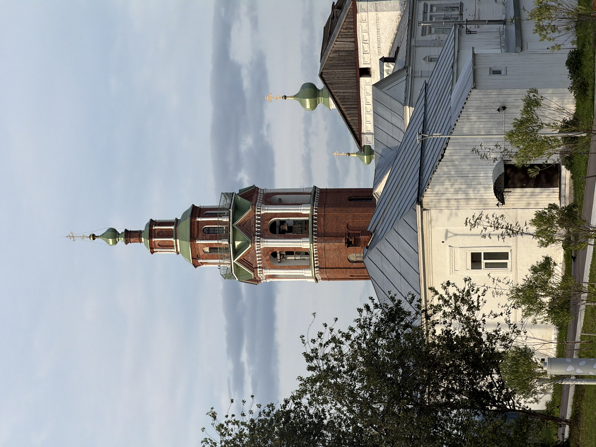 The image shows a brick bell tower with a green dome topped by a golden cross, characteristic of Russian Orthodox churches. The tower is partially obscured by a white building with a metal roof. In the background, another green dome with a cross is visible, suggesting the presence of a church complex. The sky is overcast with some patches of lighter clouds, indicating potentially diffused daylight. There are trees and green grass in the foreground and to the right of the buildings. The scene appears to be an exterior view of a historical or religious site. There is no visible text in the image. The orientation of the image suggests it was taken in portrait mode. The time of day is likely daytime, given the lighting.