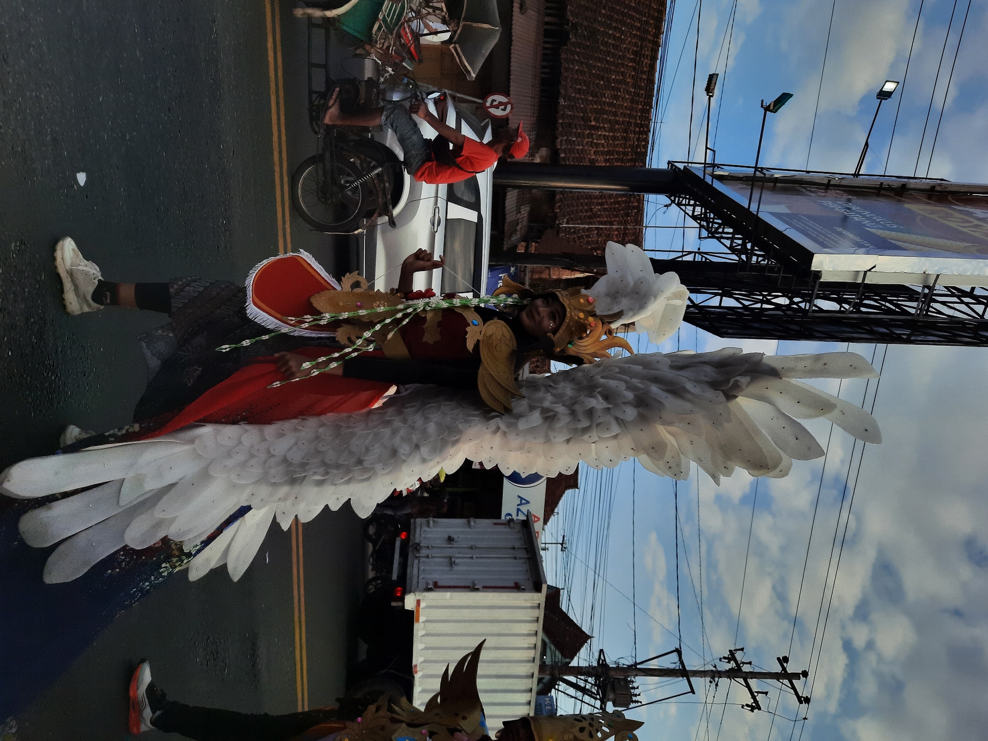 In Kutoarjo, Indonesia, on a paved street under a cloudy sky, a person in a large, ornate costume with white feathered wings walks forward. The costume features a golden headdress, a red cape, and intricate gold detailing on the chest. The person is wearing white sneakers and black leggings. In the background, a man on a motorcycle with a sidecar, wearing a red shirt and a red cap, rides past. A silver car is also visible, along with buildings with traditional tiled roofs and a large billboard. Further back, a white shipping container truck is parked. The scene appears to be a street procession or parade.
