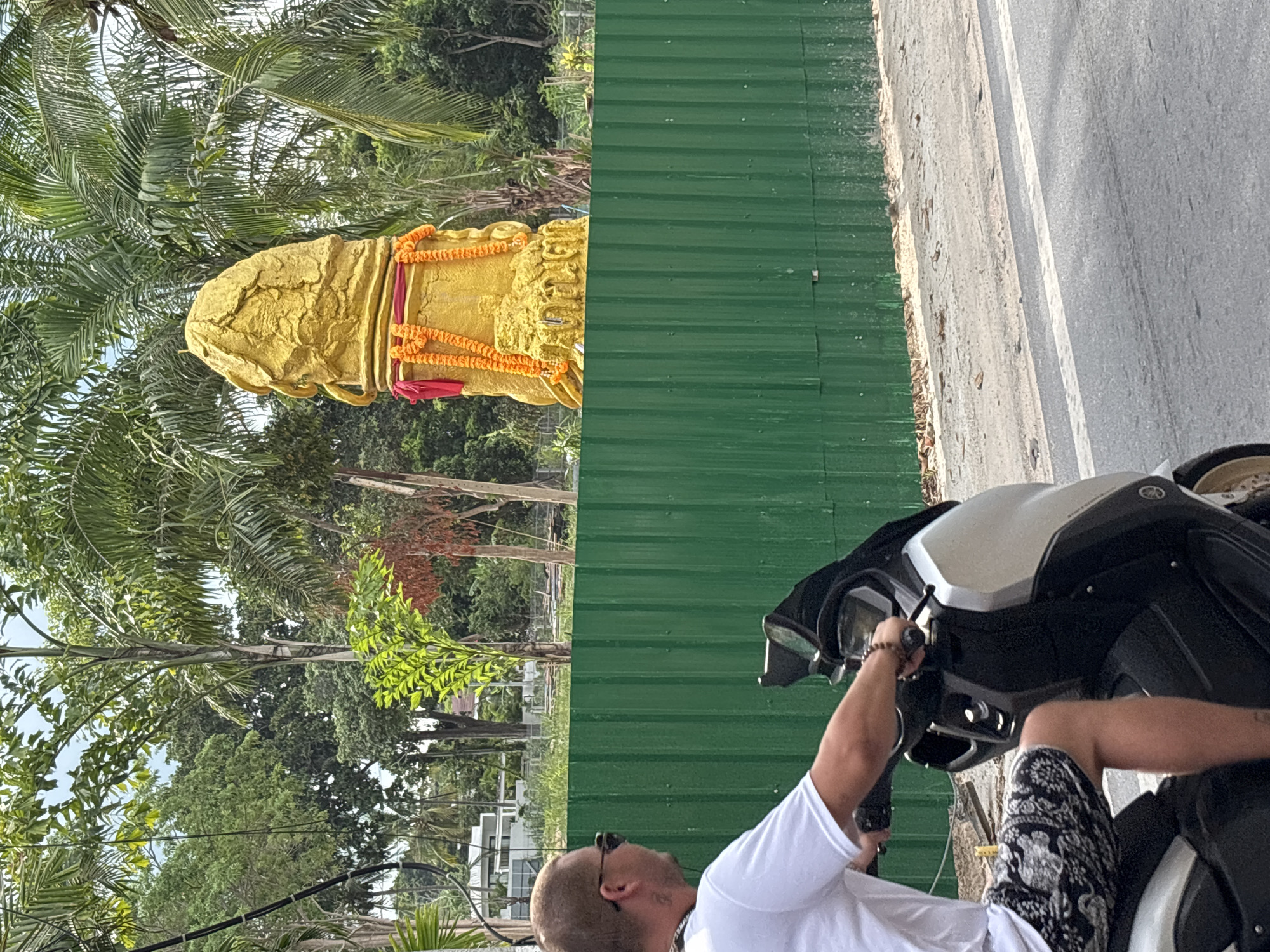 The image shows a man sitting on a scooter in บ้านบางรักษ์, Thailand, near a street with a green corrugated metal fence. A golden phallic-shaped statue is adorned with flower garlands. The background includes trees, including palm trees.