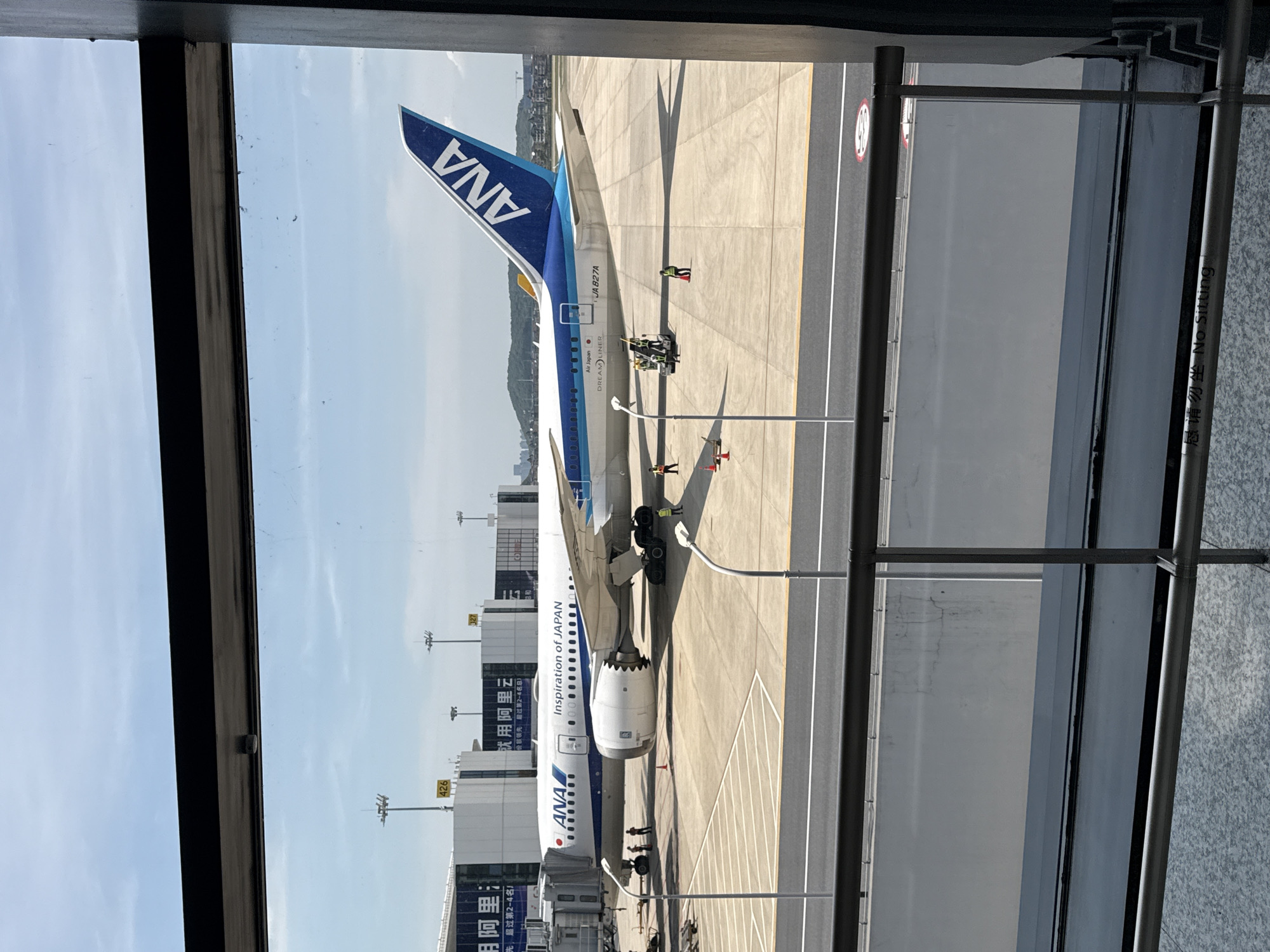 The image shows an ANA (All Nippon Airways) airplane parked at an airport gate. The plane is a Boeing 787 Dreamliner, as indicated by the "DREAMLINER" text on its fuselage. The aircraft's registration number is JA827A, and it displays the slogan "Inspiration of JAPAN". The airplane is connected to a jet bridge, suggesting passengers may be boarding or disembarking. Several airport workers are visible on the tarmac around the plane, some in high-visibility vests, possibly engaged in ground operations. The airport terminal building is visible in the background, with signage in Chinese. The scene takes place at Xiaoshan International Airport (Hangzhou Xiaoshan International Airport), located in the Xiaoshan District of Hangzhou, China. The environment is an outdoor airport apron on a clear day, with a blue sky and some clouds. The view is framed by a dark window frame, suggesting the photo was taken from inside a building, possibly a terminal or a waiting area.