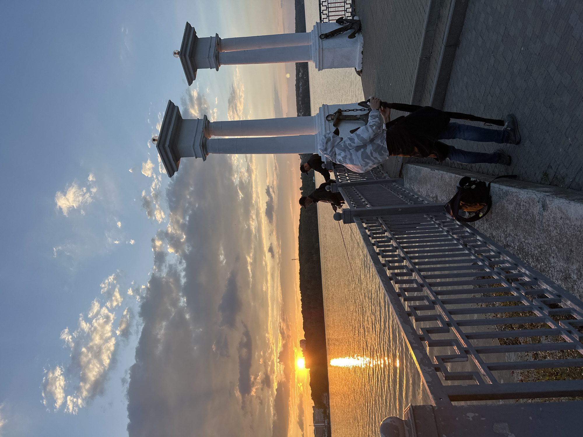 The image captures a sunset over the Ternopil pond in Ternopil, Ukraine. A picturesque scene unfolds with a body of water reflecting the golden hues of the setting sun, casting a warm glow on the clouds. In the foreground, a person in a camouflage hoodie stands holding a large umbrella, their back to the camera. Beside them, a backpack rests on a concrete ledge.
Further along the waterfront, two individuals are engaged in fishing, their fishing rods extended towards the water. The background reveals a lush green shoreline dotted with buildings, adding a touch of urban landscape to the natural beauty. Two prominent white columns with ornate tops stand on the promenade, lending a classical architectural element to the scene. Anchors are visible at the base of the columns, hinting at a maritime connection. The overall atmosphere is serene, with a sense of calm as the day draws to a close.