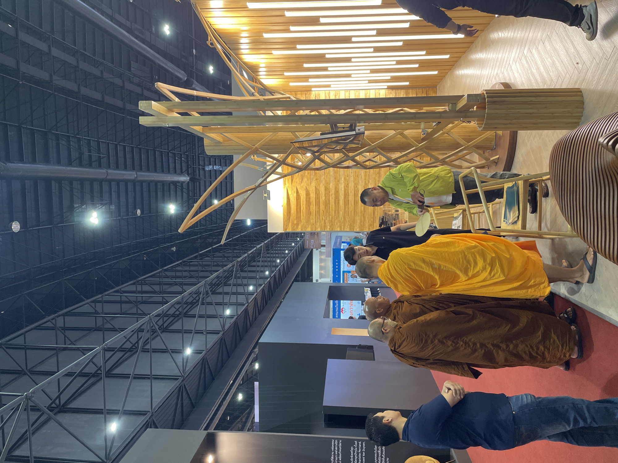 This image depicts an indoor scene, likely an exhibition or trade show, held in Pak Kret City Municipality, Thailand. The setting features a high, dark industrial-style ceiling with exposed metal girders, support beams, and ductwork, illuminated by numerous bright spotlights.
A prominent feature is an intricate, curved wooden structure, possibly an art installation or display booth, brightly lit from below by linear LED lights. Adjacent to this, various wooden furniture pieces, including a unique slatted design, are visible.
Several people are present, including three men wearing traditional Buddhist robes—two in brown and one in a vibrant yellow—standing on a red carpeted area. A man in a lime green shirt is seated nearby, seemingly interacting with an exhibit or demonstrating something on a wooden counter. Another man in a blue shirt and jeans stands on the red carpet, looking upwards.
The activity appears to be people observing or participating in an exhibition, with the monks possibly visiting or attending a special event. The overall ambiance is well-lit and attentive.
Visible text in Thai discusses concepts related to "architects," "furniture," and "design," confirming the exhibition's theme. English names such as "Charlotte Perriand" and "Vincent Van Duysen" are also discernible, suggesting design influences or featured artists/designers. A sign displaying "PAT F103" is also visible.