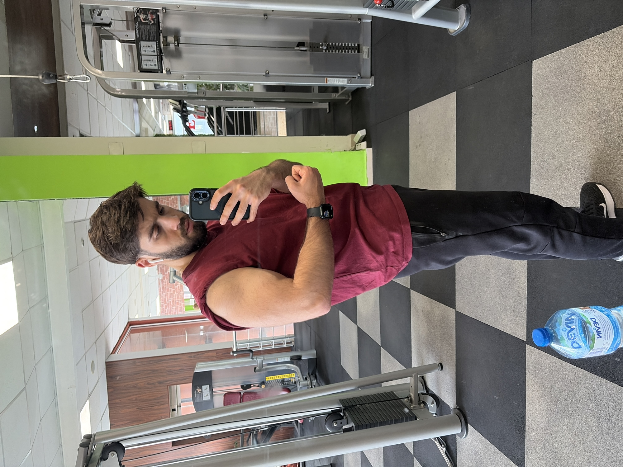A man is standing in a gym, taking a selfie to showcase his bicep. He has a beard and is wearing a maroon t-shirt, black sweatpants, and a smartwatch. He holds a smartphone in his left hand. The gym has a black and white checkered floor and various exercise machines. A bottle of "Devin" water sits on the floor near his feet, indicating the location is likely in Bulgaria, as "Devin" is a popular Bulgarian brand. The specific city is Sofia, Bulgaria.