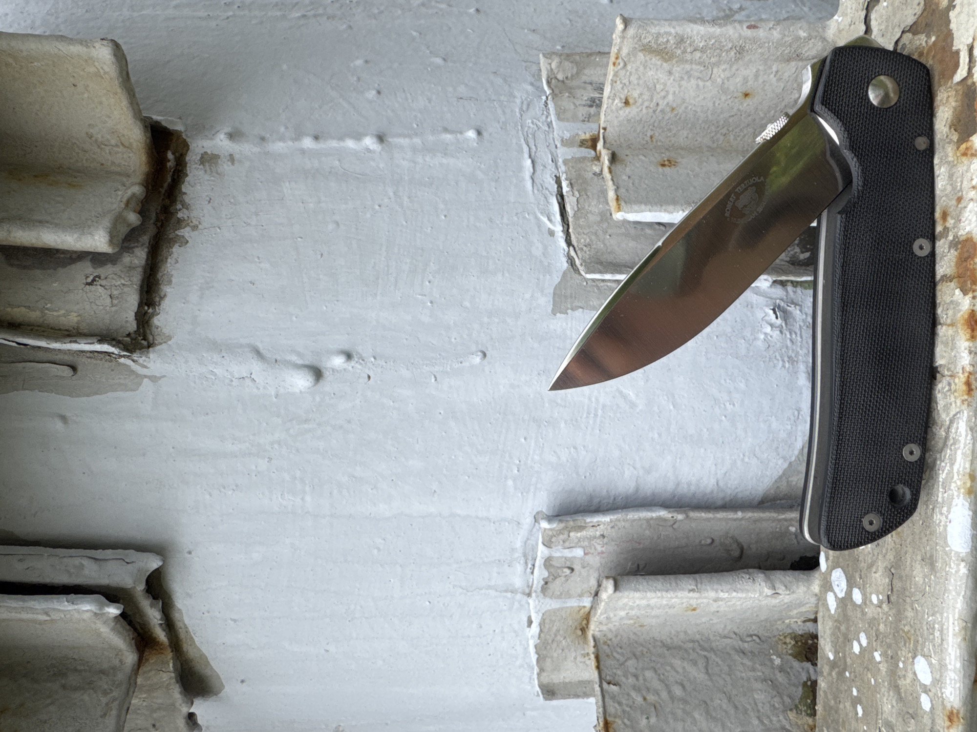 A bright, well-lit close-up shot features a partially open folding knife positioned vertically against a textured, light-colored metal structure. The knife has a black, textured handle and a reflective silver blade, which points slightly upwards and to the left. Engraved on the blade, the text "ROBERT TERZUOLA" is clearly visible, along with a bear's head logo and the word "FALCON GEAR" below it. The knife appears to be held in place against the structure, possibly wedged or resting on a protrusion.
The immediate surroundings consist of painted metal structural elements, likely beams or supports, characterized by peeling light gray or off-white paint, visible rust, and rough textures. These elements show signs of age and wear, with exposed metal and corrosion in the joints and crevices. The background is a similarly textured, painted surface, possibly a wall or another large structural component. Small, raised bumps or old weld marks are scattered across the painted surfaces. The bright, even lighting suggests it is daytime. No people or dynamic activity are depicted. The scene is static, focusing on the knife within its weathered, industrial or architectural setting, possibly in Moscow, Russia.