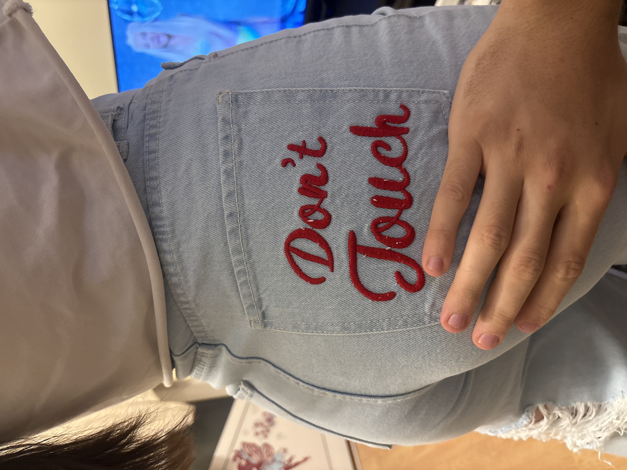 The image shows the rear of a person wearing light blue denim jeans. The right back pocket of the jeans has the embroidered text "Don't Touch" in red. A person's hand rests on the right back pocket, partially obscuring the text. The jeans appear distressed with frayed edges around the lower leg. The background is blurred and includes glimpses of a white garment, a TV screen, and a table with a floral pattern. The scene does not provide enough detail to determine a location in Burgas, Bulgaria.
