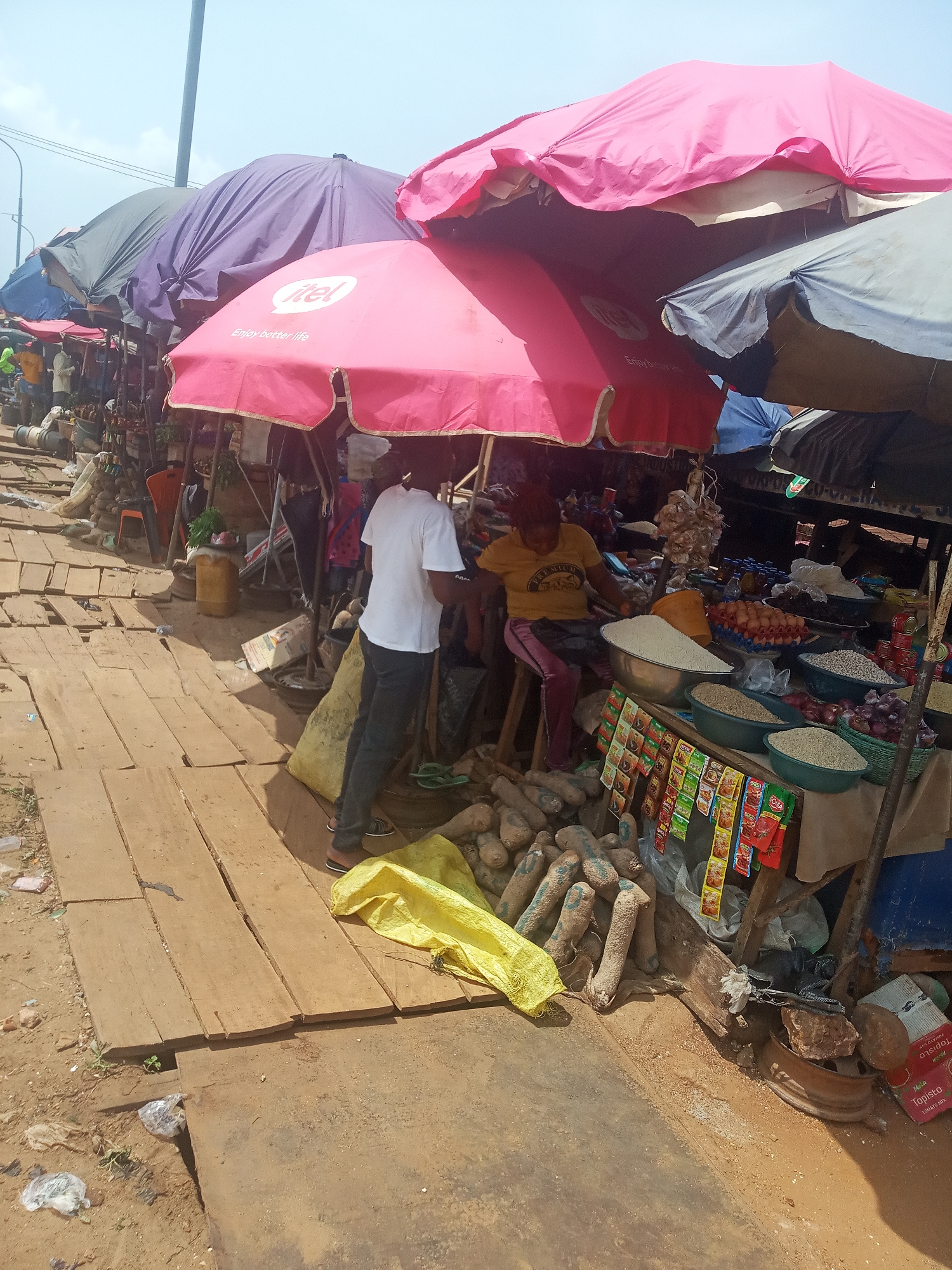 This image captures an outdoor market scene in Nsugbe, Nigeria, under a bright, possibly partly cloudy, daytime sky. The setting features numerous market stalls, primarily covered by large, colorful umbrellas providing shade. The ground is a mix of bare earth and wooden planks laid out to form walkways.
In the foreground, a market stall is actively tended. A person in a white t-shirt and dark trousers stands facing the stall, their back mostly to the camera. Adjacent to them, a vendor wearing a yellow t-shirt with "PREMIUM" partially visible and pink/purple trousers is seated on a stool behind the stall, overseeing the goods.
The stall displays a variety of fresh produce and packaged goods. Large quantities of yams or similar tubers are piled on the ground to the right. On the vendor's table, several bowls hold different grains and beans, along with trays of eggs and baskets of onions. Garlands of garlic hang above the produce. Various small sachets of spices and seasonings, including "ROYCO," "KNORR," and "SOLA" brands, are neatly hung from the front of the stall. A cardboard box labeled "Topista Tomato Mix" is also visible near the ground.
The most prominent umbrella, colored pink, displays the text "itel Enjoy better life," suggesting a brand sponsorship for the market vendors. Other umbrellas are purple, grey, and blue, stretching into the background where more market activity and figures are faintly visible. The scene depicts a typical bustling day of commerce in an African market.