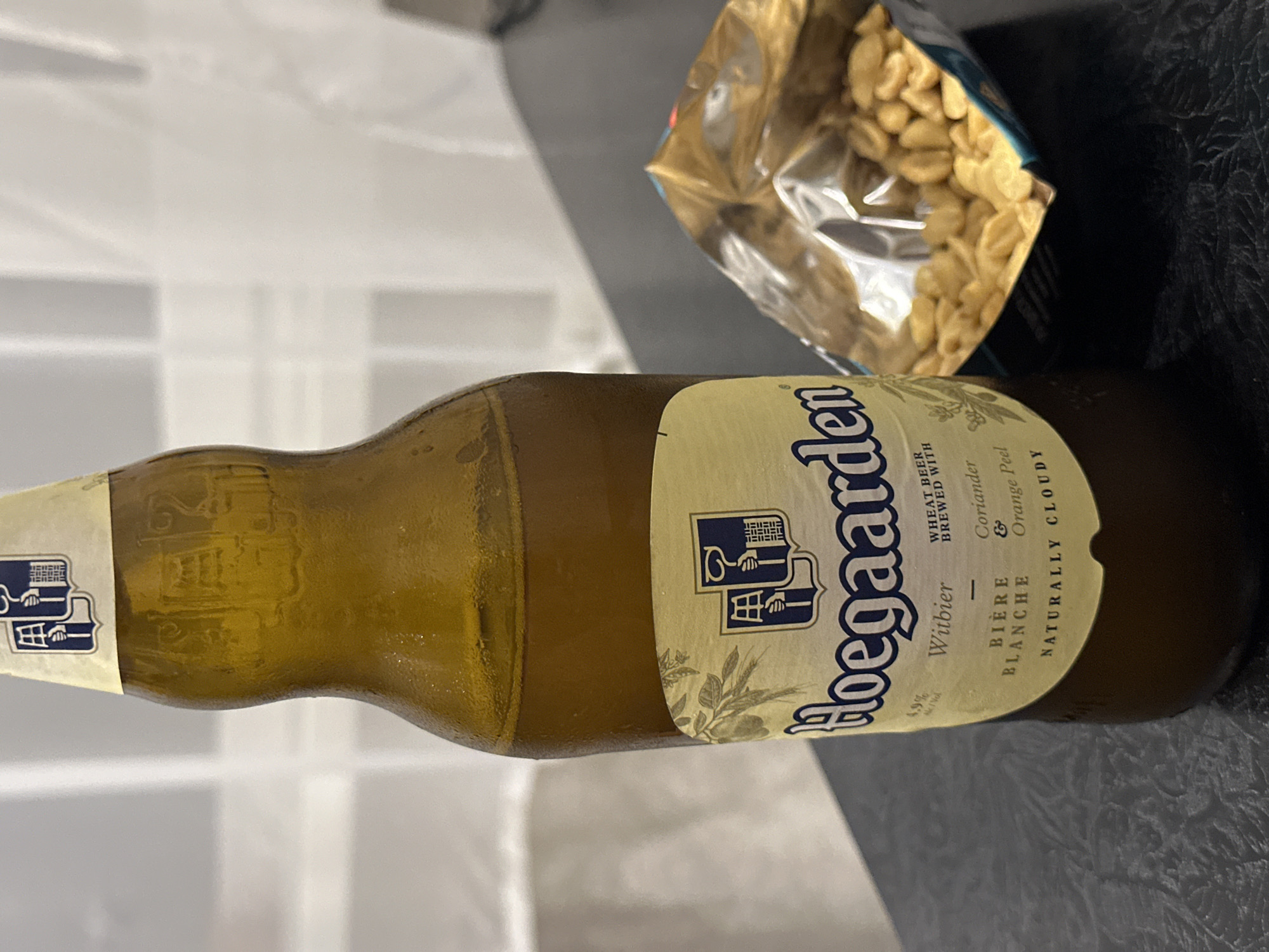 A close-up shot focuses on a brown glass bottle of Hoegaarden Witbier, condensation on its surface, placed on a dark textured surface, likely a table. The beer bottle's label, featuring the brand name "Hoegaarden" in blue lettering and a shield emblem, is clearly visible. To the right of the bottle, a partially opened bag of peanuts spills onto the surface. The background is blurred, showing a white curtain or blind. The image does not provide information about the city of Energetik, Russia, or any individuals.