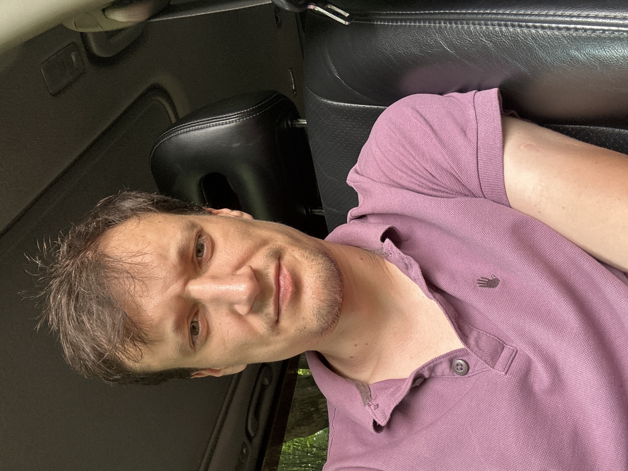 A close-up, front-facing selfie of a man sitting in the driver's seat of a car. The man has brown hair, a light complexion, and is wearing a purple polo shirt. The car's black leather interior is visible, including the headrest and part of the dashboard. The background shows blurred green foliage, suggesting the car is parked outdoors. The man's expression is neutral. This image was taken in Shatava, Ukraine.