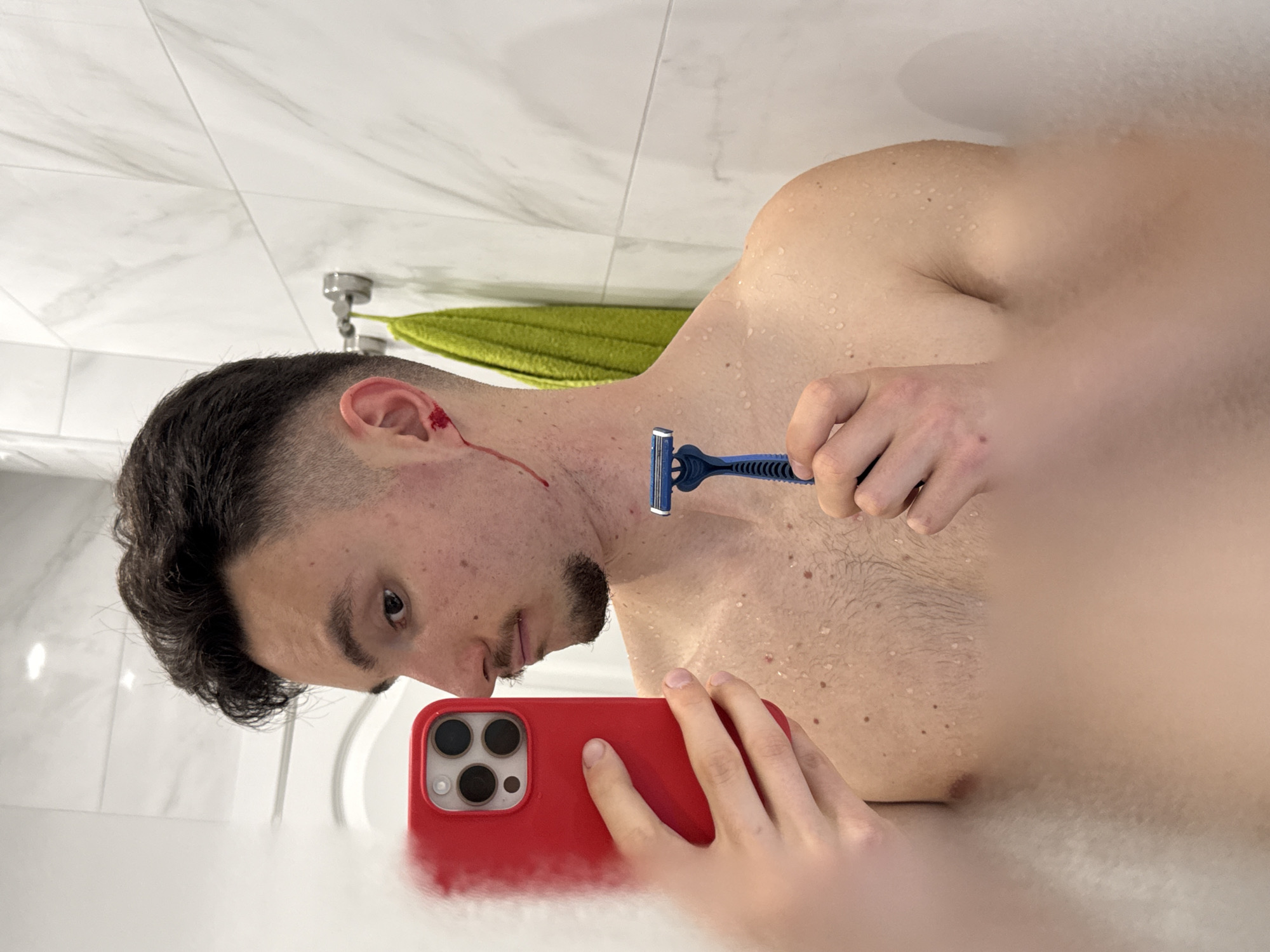 A shirtless man is in a bathroom, holding a razor to his neck in one hand and a red smartphone in the other, presumably taking a selfie. He has a small cut on his ear with a trickle of blood running down his neck. The bathroom has white marble-like tiled walls and a green towel hanging on a silver towel rack. The location of the image is not possible to determine based on the provided visual information.