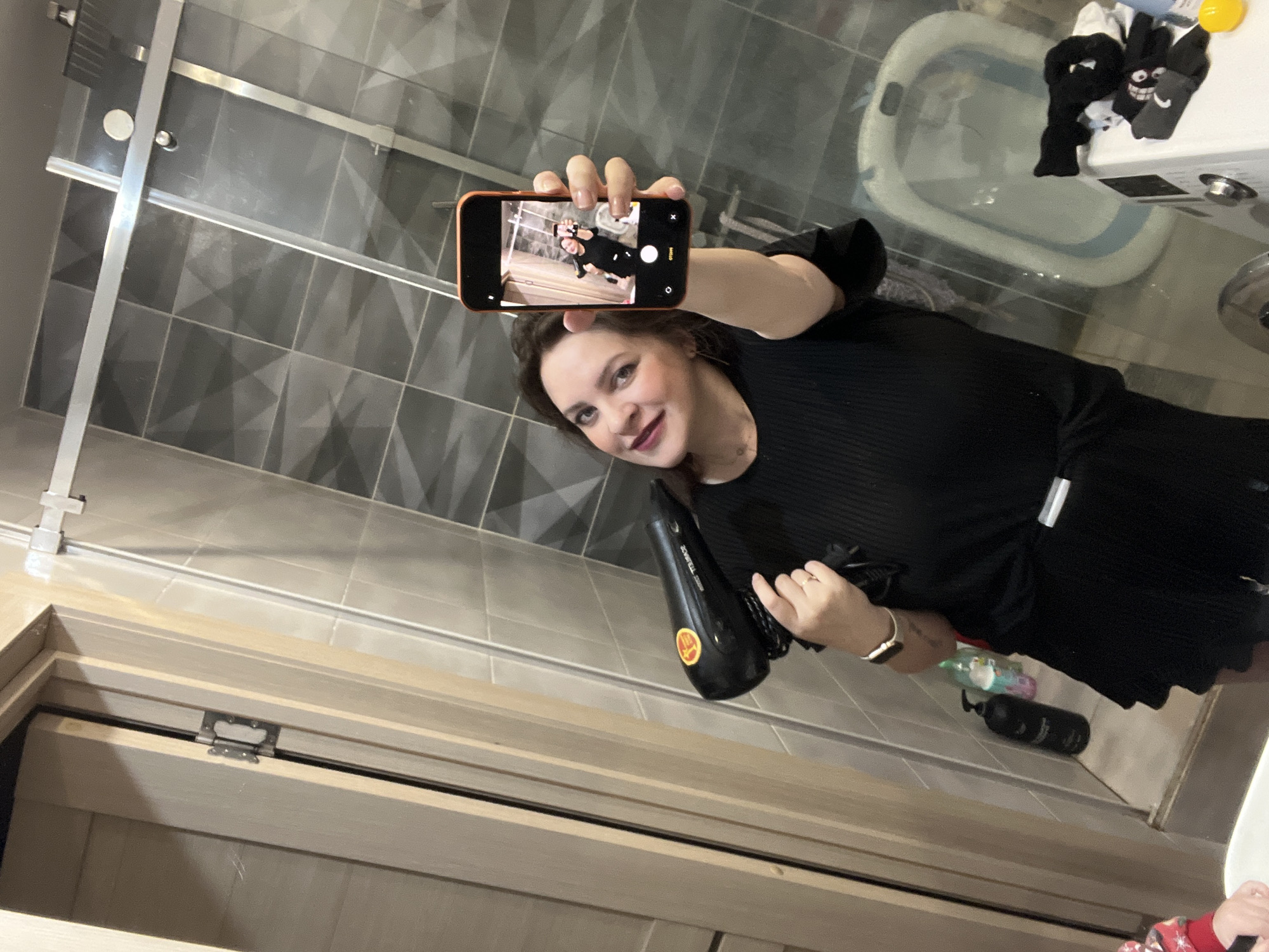 A middle-aged woman with dark hair is taking a selfie in a bathroom. She is holding a cell phone in her right hand, and the phone is displaying a smaller version of the same image. She is also holding a black hair dryer in her left hand. She is wearing a black dress.
The bathroom has gray and white tiled walls and floor. A bathtub with some items on it is visible in the background. The location is Noviy, Russia.