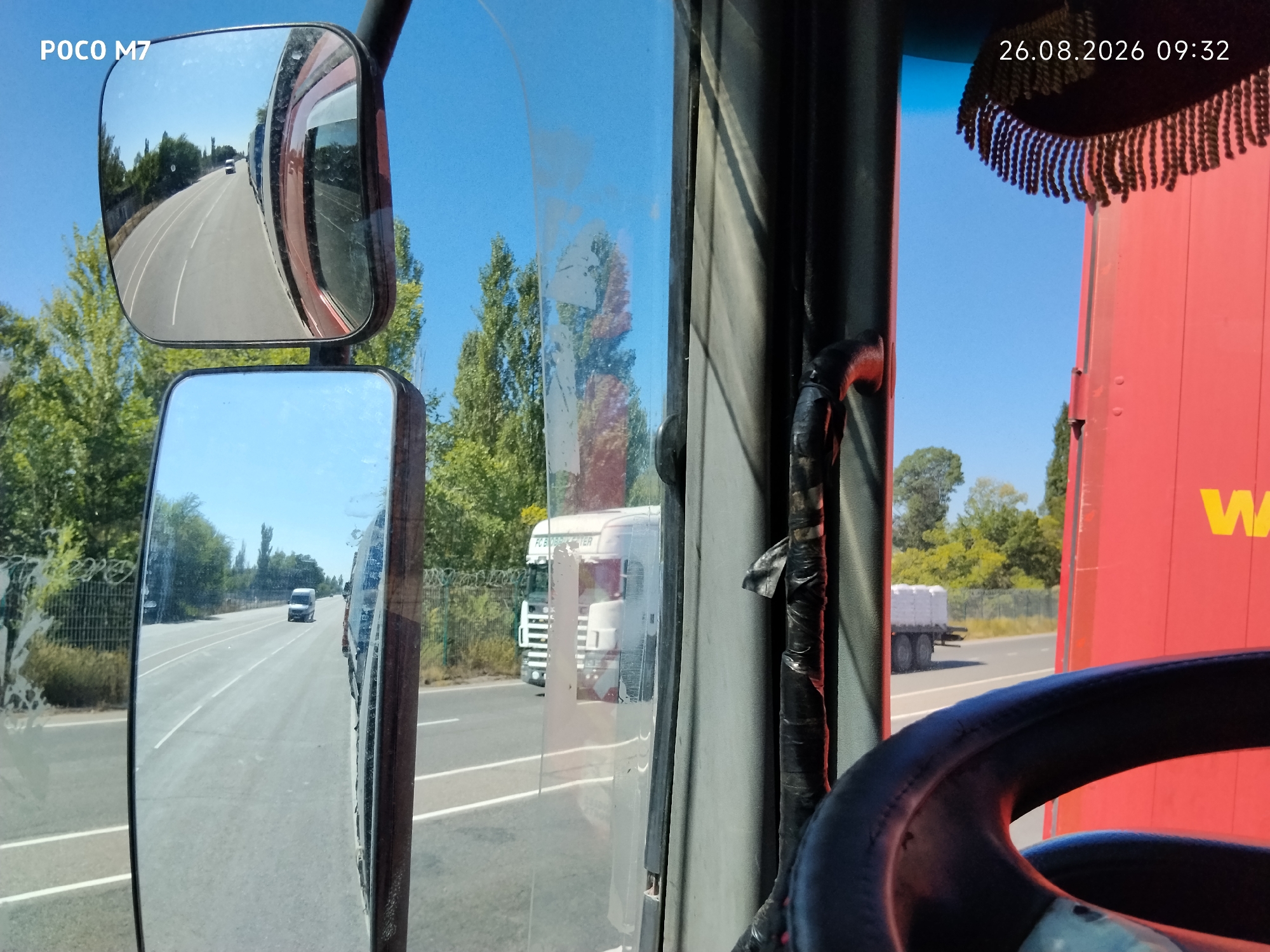 The image is taken from the interior of a vehicle, likely a truck, looking out through the windshield and mirrors. A date and time stamp indicates "26.08.2026 09:32". The driver's side mirror reflects a road with passing trees and another vehicle in the distance. A smaller secondary mirror is also visible. The view through the windshield shows a road with passing trees and another truck. To the right, a large red surface is visible, possibly the side of a trailer or a building, with a yellow "W" logo. The scene appears to be on a sunny day. The location is identified as Ауқатты, Kazakhstan.