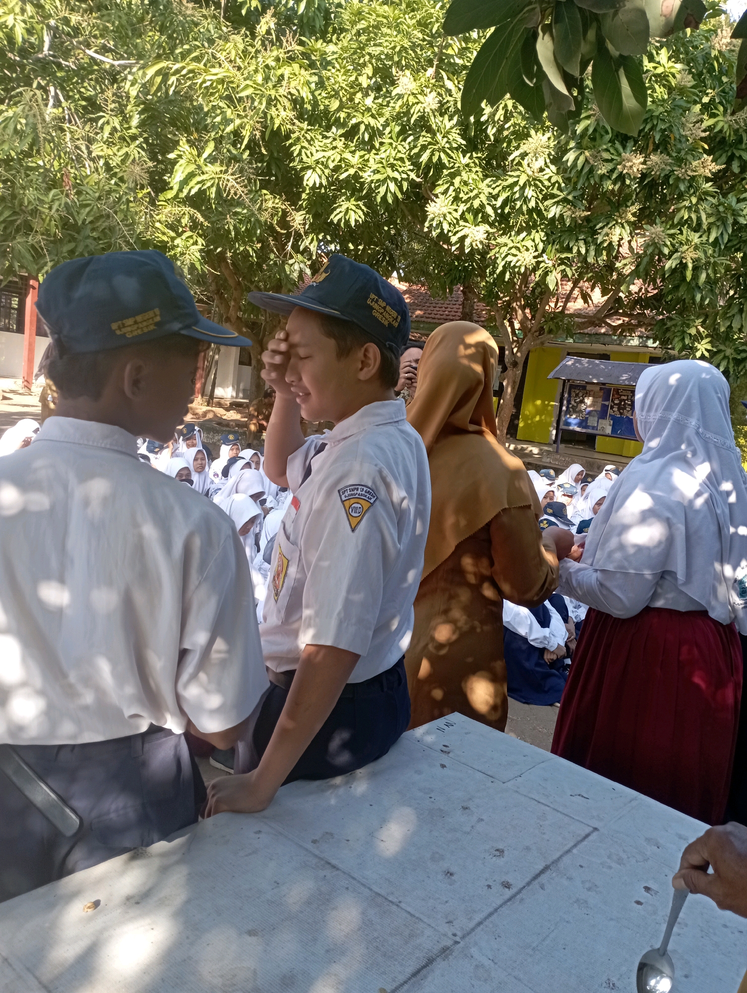 The image depicts a sunny outdoor scene, likely at a school in Banyuurip, Indonesia. In the foreground, two male students in white uniforms and dark caps face each other. The student on the right, with his eyes closed and hand on his forehead, appears to be looking away from the other student. The student on the left is partially obscured, facing away from the camera and toward the other student. Behind them, a group of students, predominantly female wearing white headscarves and uniforms, are gathered. One female student in a brown hijab and another in a white hijab are visible between the foreground students and the larger group. The background features lush green trees, suggesting a campus or park setting. A yellow structure with a bulletin board is visible to the right. The lighting indicates a bright, sunny day, casting distinct shadows.