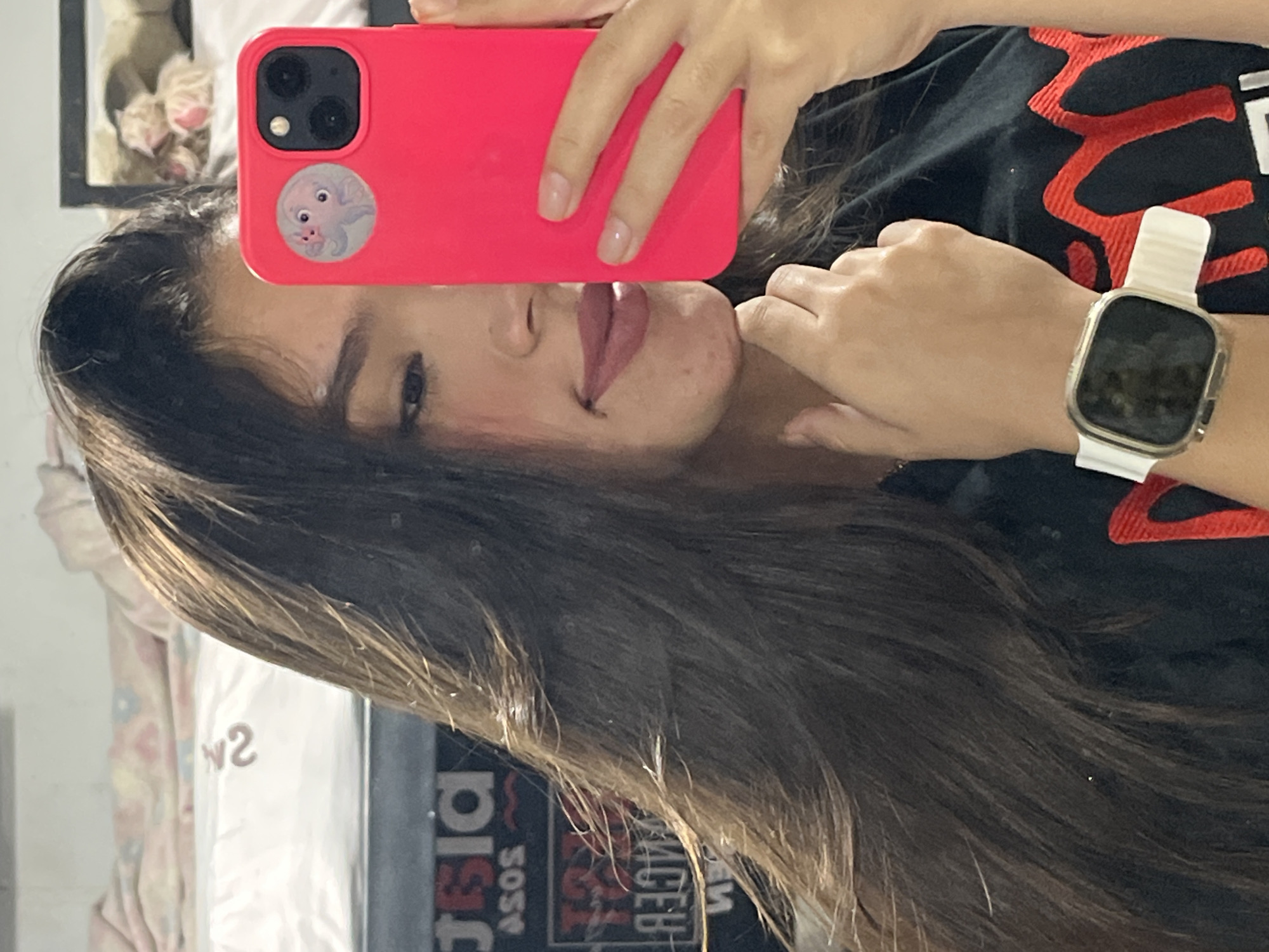 The image shows a young woman taking a selfie with her smartphone. She is wearing a black t-shirt with a red design. On her left wrist, she is wearing a white smartwatch. The background appears to be an indoor setting with a bed and some posters or signs on the wall. One sign has the text "ASOS" visible. The overall lighting suggests an indoor environment, possibly a bedroom.