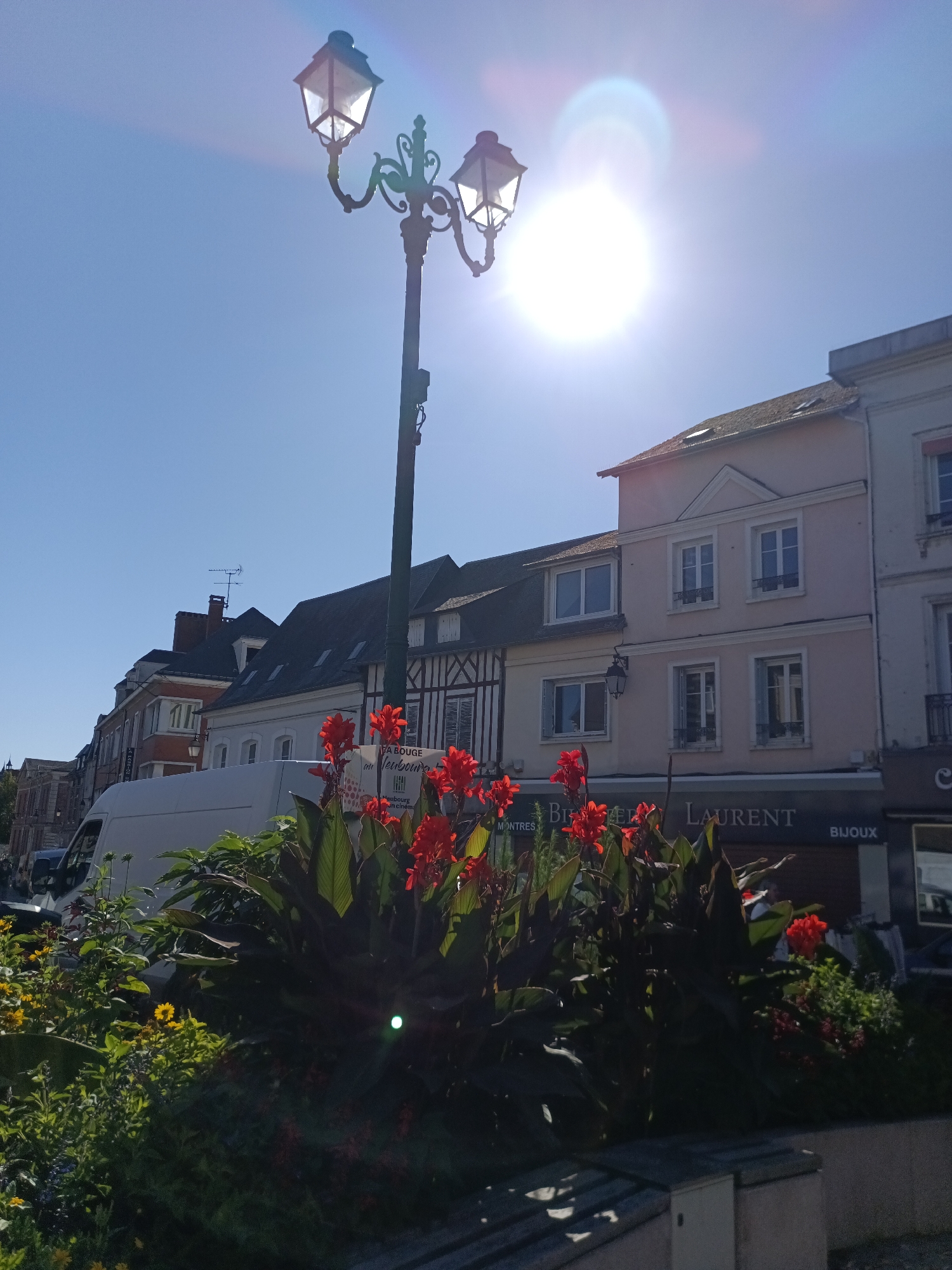 The image depicts a street scene in Le Neubourg, France, on a clear, sunny day. In the foreground, a cluster of vibrant red flowers and lush green foliage is prominent. Behind the flowers, a white van is parked. Further back, a row of traditional French buildings lines the street. These buildings feature multi-story structures with pitched roofs, some with visible dormer windows. The architecture includes elements of half-timbered construction on one building. A decorative street lamp with multiple light fixtures stands tall on the left side of the frame, silhouetted against the bright sky. The sun is high in the sky, causing lens flare and making it difficult to discern details in the brightest areas. A shop sign reading "BI BIEREN LAURENT" and "BIJOUX" is visible above a storefront. The overall impression is a picturesque European town square or street.