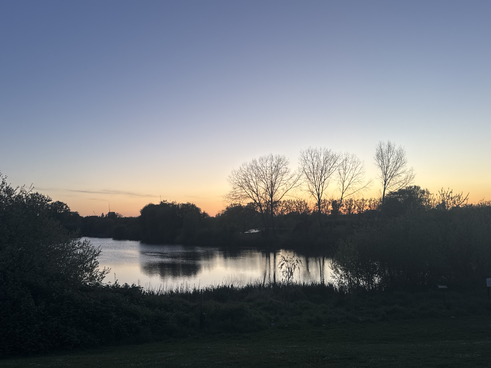 This wide shot captures a tranquil landscape at dusk, likely in a natural area within London, United Kingdom. The sky, dominating the upper half of the image, displays a beautiful gradient, shifting from a deep indigo blue at the top through soft purples and lavenders, to a warm orange-yellow glow along the horizon line. This bright band of light indicates either sunset or sunrise, with the muted, darkening foreground suggesting sunset.
A calm body of water, resembling a lake or wide river, occupies the mid-ground, serving as a reflective surface for the changing sky colors and the dark forms of the trees and bushes along its banks. Several prominent deciduous trees, silhouetted against the brightest part of the horizon, stand bare, their intricate branches clearly visible. Other trees and dense bushes form dark masses along the water's edge, framing the scene.
The immediate foreground consists of shadowy land covered in grass and low-lying vegetation. In the far distance on the left horizon, subtle silhouettes of what appear to be cranes or urban structures can be seen, hinting at the proximity of a city. No specific human activity is taking place. The overall atmosphere is peaceful and serene, emphasizing the natural beauty of the transition from day to night. On the far right bank, a white sign with dark, illegible text is visible.