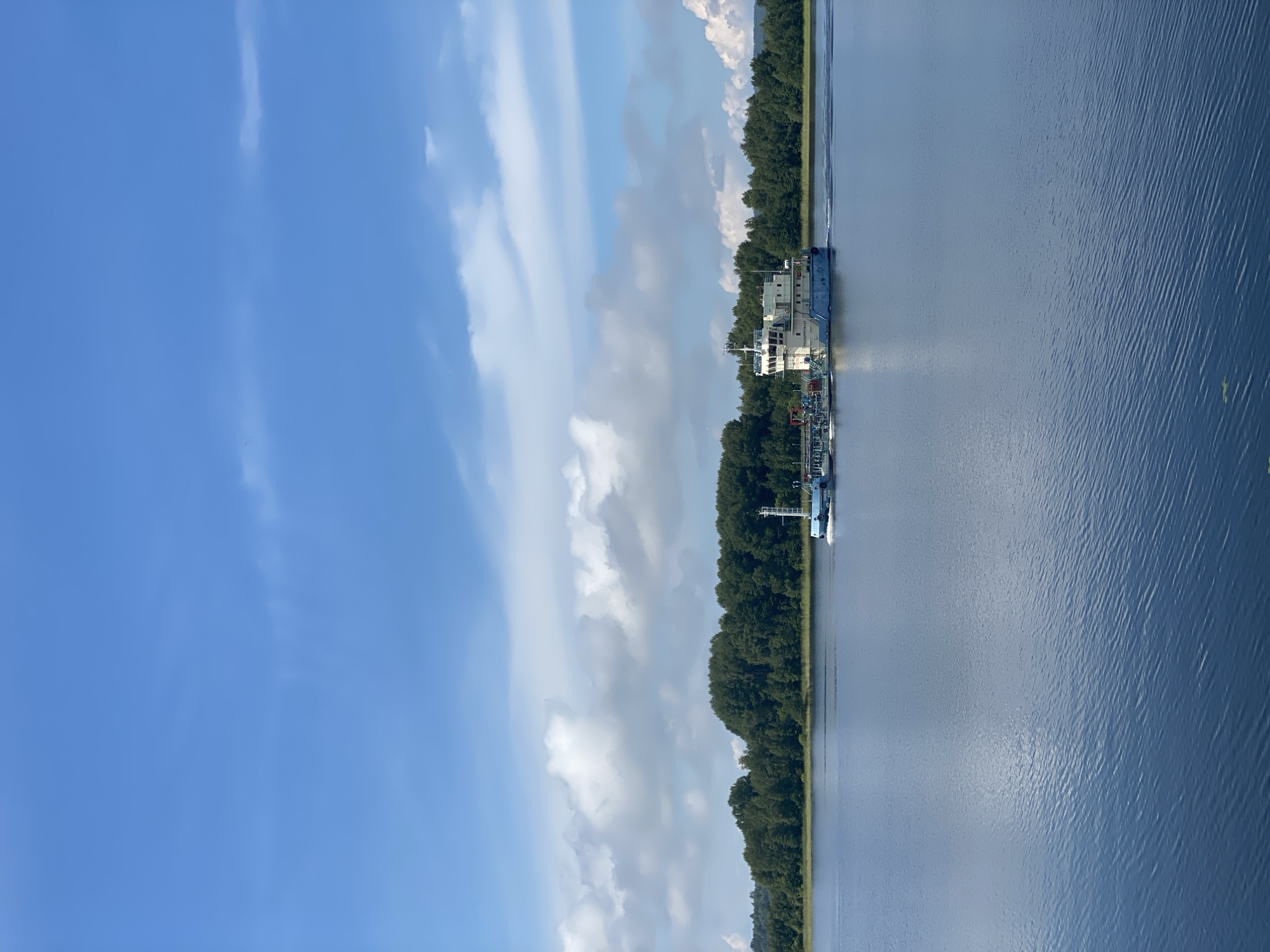 A barge moves across a calm, blue body of water under a bright blue sky with scattered white clouds. The barge, painted primarily white and light blue, has a white superstructure with multiple windows and a navigation mast. It appears to be carrying cargo, with visible pipes and tanks on its deck. In the background, a dense line of dark green trees forms a horizon along the far shore. The water reflects the sky and trees, creating a serene natural landscape. The scene is likely a river or lake in Boyarshchina, Russia, given the presence of the barge and the forested environment. There are no people visible in the image.