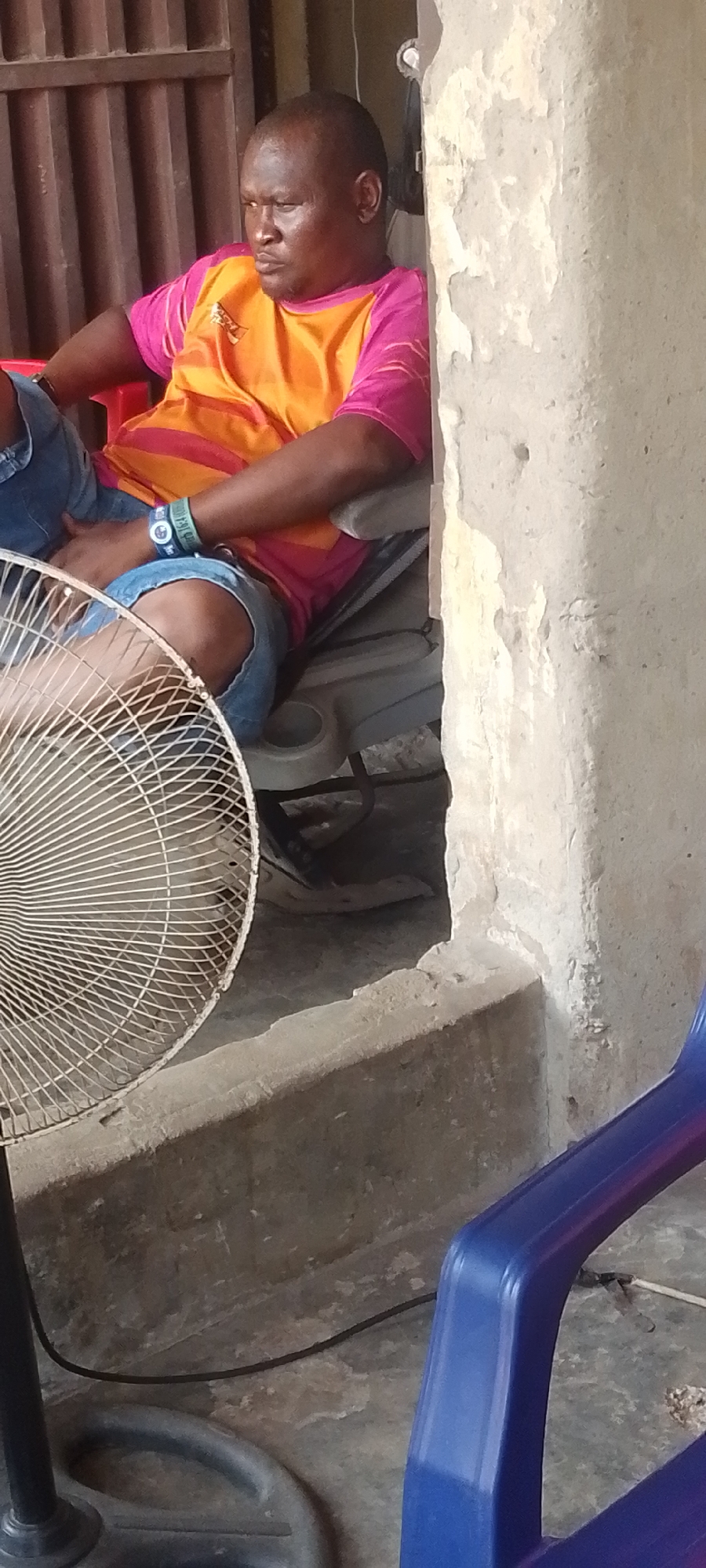 In Lokoja, Nigeria, an adult man with a shaved head is seated in a red plastic chair, wearing a pink and orange t-shirt and blue jean shorts. A watch is visible on his left wrist. He sits within a concrete alcove defined by a column showing wear and deterioration. Adjacent to the alcove, vertical bars partially obscure the background. A standing fan stands to the left, and a blue plastic chair is situated to the right. The floor is concrete.
