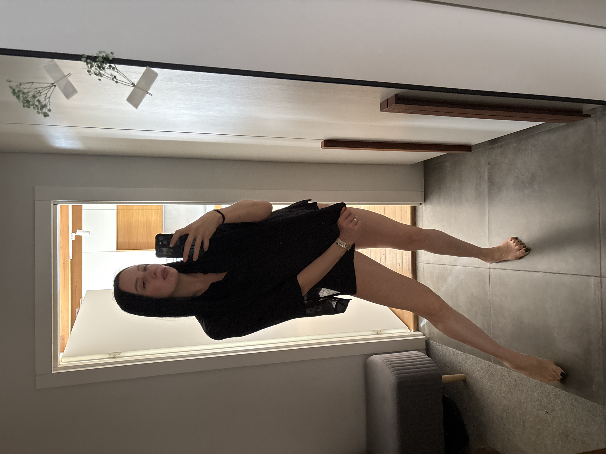 A young woman is captured in a full-length mirror selfie, standing indoors and holding a smartphone to take the picture. She has long dark hair and is wearing a dark, possibly black, short-sleeved top or dress. A gold-toned watch is visible on her left wrist, and a ring is on her left ring finger. Her toenails are painted black. She poses with her legs extended; her left foot rests on a low, raised ledge or piece of furniture, while her right foot is flat on the grey tiled floor. Her expression is neutral with a slight smile.
The setting appears to be a modern indoor space, possibly a hallway or room in Chaikovsky, Russia, with light-colored walls and grey tiled flooring. The mirror reflects a portion of the room behind her, including what looks like a doorway or window with wooden framing. Two small bunches of green foliage, resembling baby's breath, are decoratively taped to the upper section of the mirror. Above the mirror, two dark wooden rectangular pieces are mounted horizontally on the wall. A grey upholstered bench or ottoman is partially visible in the lower right corner of the image. The lighting suggests it is daytime, with light entering from the reflected background. No visible text is present in the image.