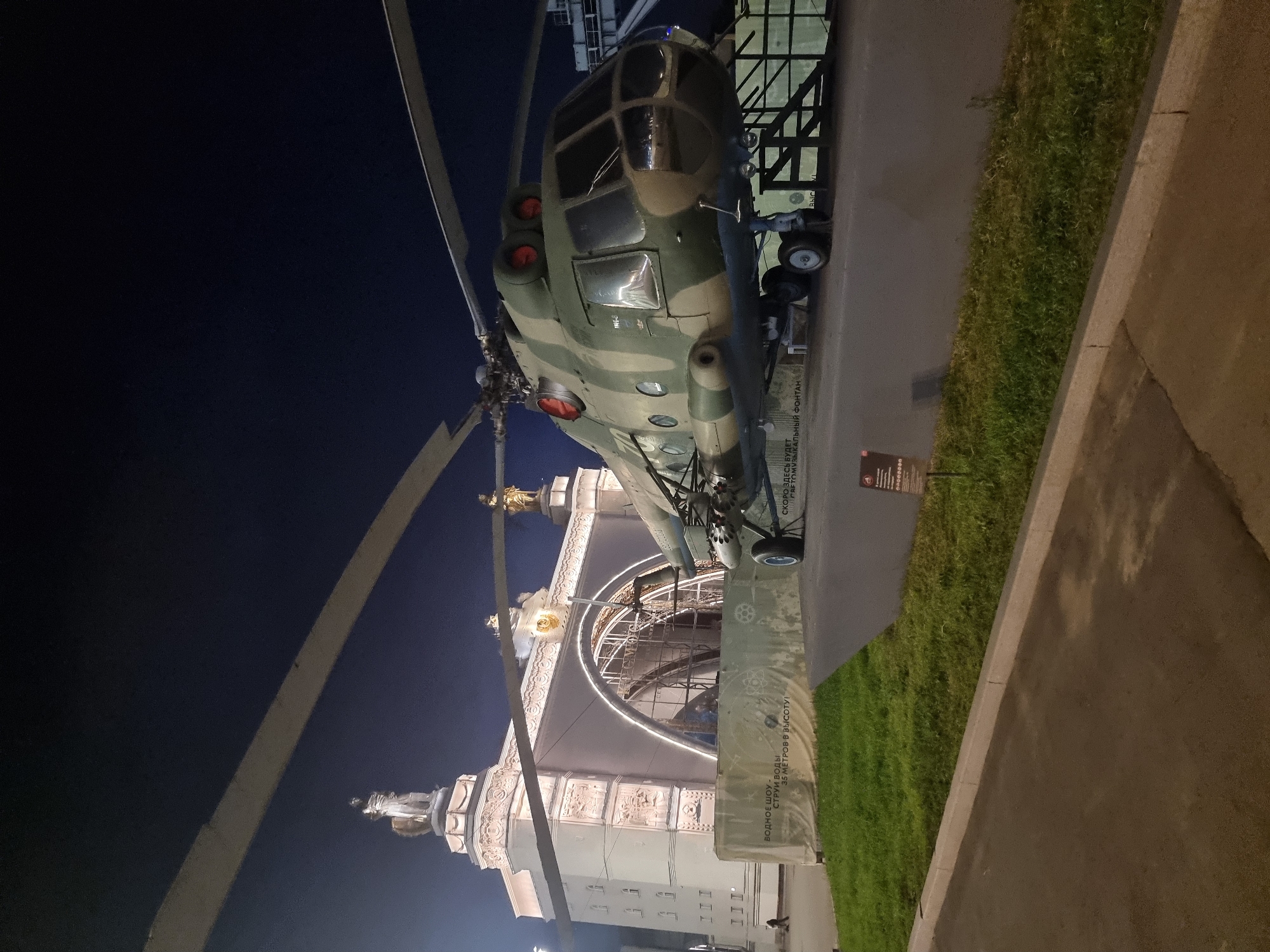 An outdoor, nighttime, low-angle shot features a camouflaged military helicopter prominently displayed on a paved area in front of a large, ornate building. The building, illuminated by numerous lights, has a grand arched entrance and is adorned with statues and decorative elements. A segment of a rotor blade extends into the upper left of the frame, with another large blade crossing the top of the image. The helicopter, a Mil Mi-8 or Mi-17 variant, is depicted with its landing gear deployed and appears to be carrying multiple rocket pods under its fuselage. In the foreground, a paved walkway runs along a strip of green grass. A light-colored fabric banner with Cyrillic text, including "ВОДНОЕ ШОУ" (Water Show) and "35 МЕТРОВ В ВЫСОТУ!" (35 meters in height!), is visible behind the helicopter, suggesting a temporary exhibition or event. Further text on the banner reads "СКОРО ЗДЕСЬ" (Soon here) and "#ТОМУЗЫКАЛЬНЫЙ ФОНТАН" (#themusicalfountain). In the far background, the dark night sky is visible. A small informational sign with a QR code is planted in the grass to the right of the helicopter. The overall scene suggests a public exhibition of military hardware in a park-like setting within Moscow, likely at the VDNKh (Exhibition of Achievements of National Economy) complex, given the architectural style and the presence of exhibition banners.