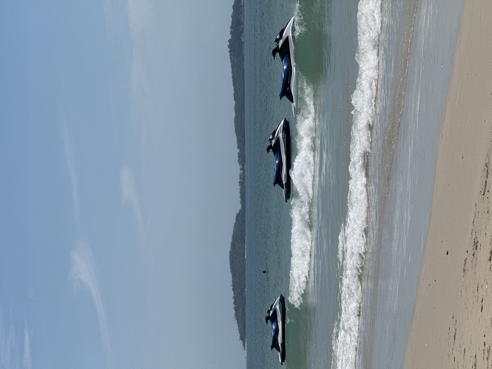 The image depicts a serene beach scene in Ko Samui District, Thailand, on a bright, clear day. Three jet skis are parked in the shallow, turquoise waters, appearing to be stationary. The nearest jet ski is on the right side of the frame, partially obscured by a gentle wave. The other two are positioned further to the left, also in the water. In the middle ground, a lone individual is swimming in the sea, their body mostly submerged. A chain of islands covered in lush green vegetation is visible in the distance, under a pale blue sky with faint wispy clouds. The foreground features a sandy beach with a light scattering of small dark pebbles. The waves are gently rolling onto the shore, creating a foamy white crest.