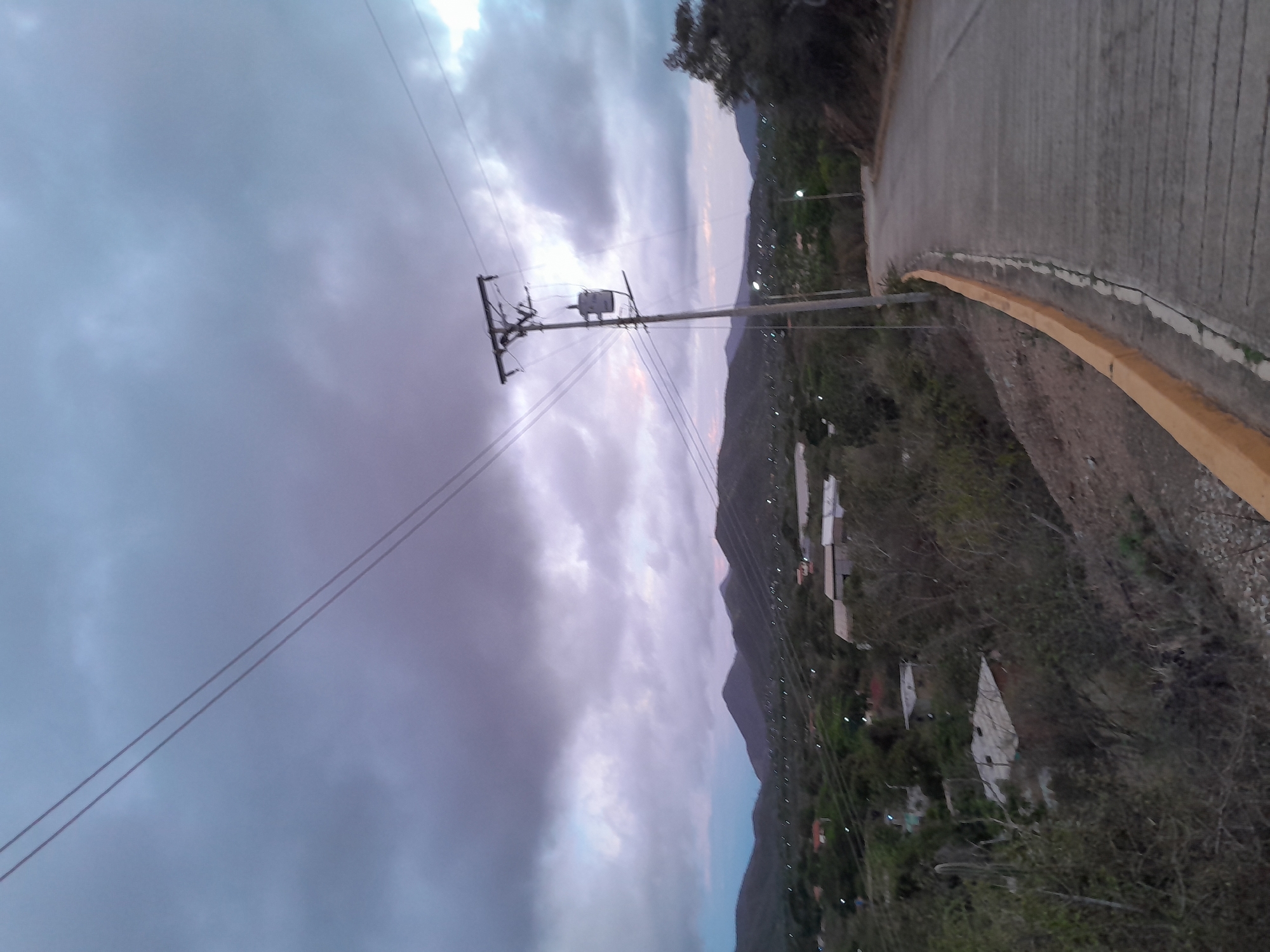 A cloudy, overcast sky looms over a rural landscape with rolling hills in the distance. A paved road curves away to the right, bordered by a yellow curb and dry, scrubby vegetation. A wooden utility pole with a transformer and wires stands on the left side of the road. In the middle ground, a scattering of small houses and buildings are visible amidst trees and foliage. The scene suggests a remote, possibly underdeveloped area, with indications of electrical infrastructure connecting the scattered dwellings. The overall mood is subdued, likely due to the heavy cloud cover and the twilight hour.