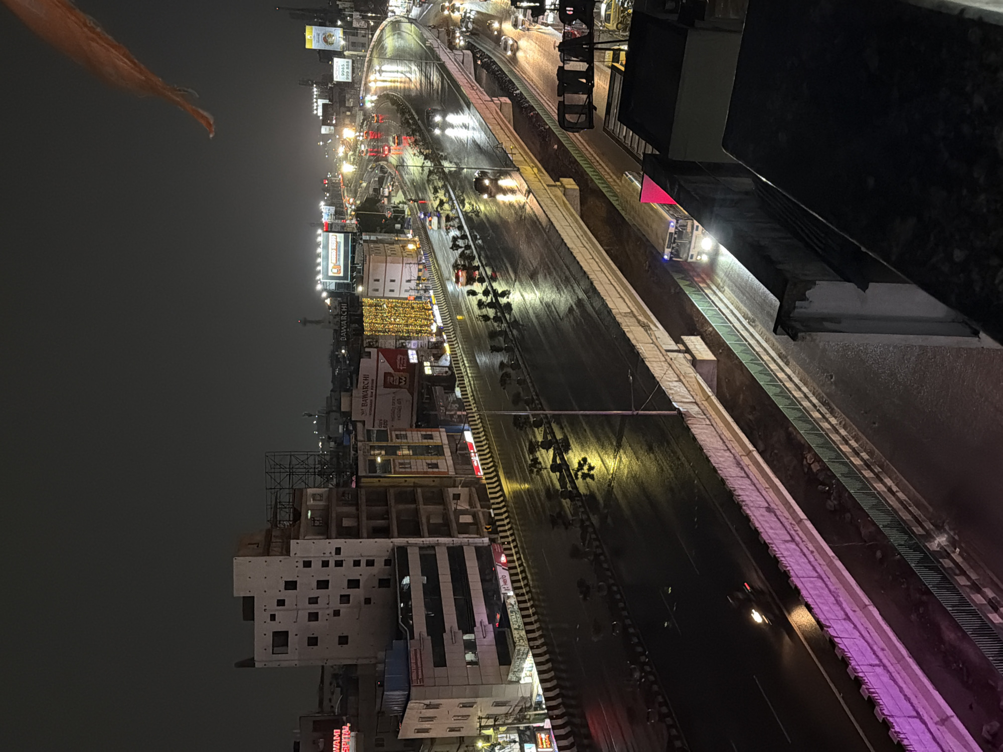 This image captures a nighttime view of a multi-level elevated highway in Shamshabad, India, likely during or shortly after rainfall. The wet asphalt reflects the bright headlights of numerous vehicles, including cars, trucks, and a bus prominently visible in the foreground. Buildings line the sides of the highway, some adorned with illuminated signage and decorative lights. One large billboard displays the word "BAWARCHI" in illuminated letters. The sky is dark and overcast. A distinctive orange flag is visible in the upper right corner of the frame. The scene depicts a busy urban environment with active traffic flow despite the nighttime and wet conditions.