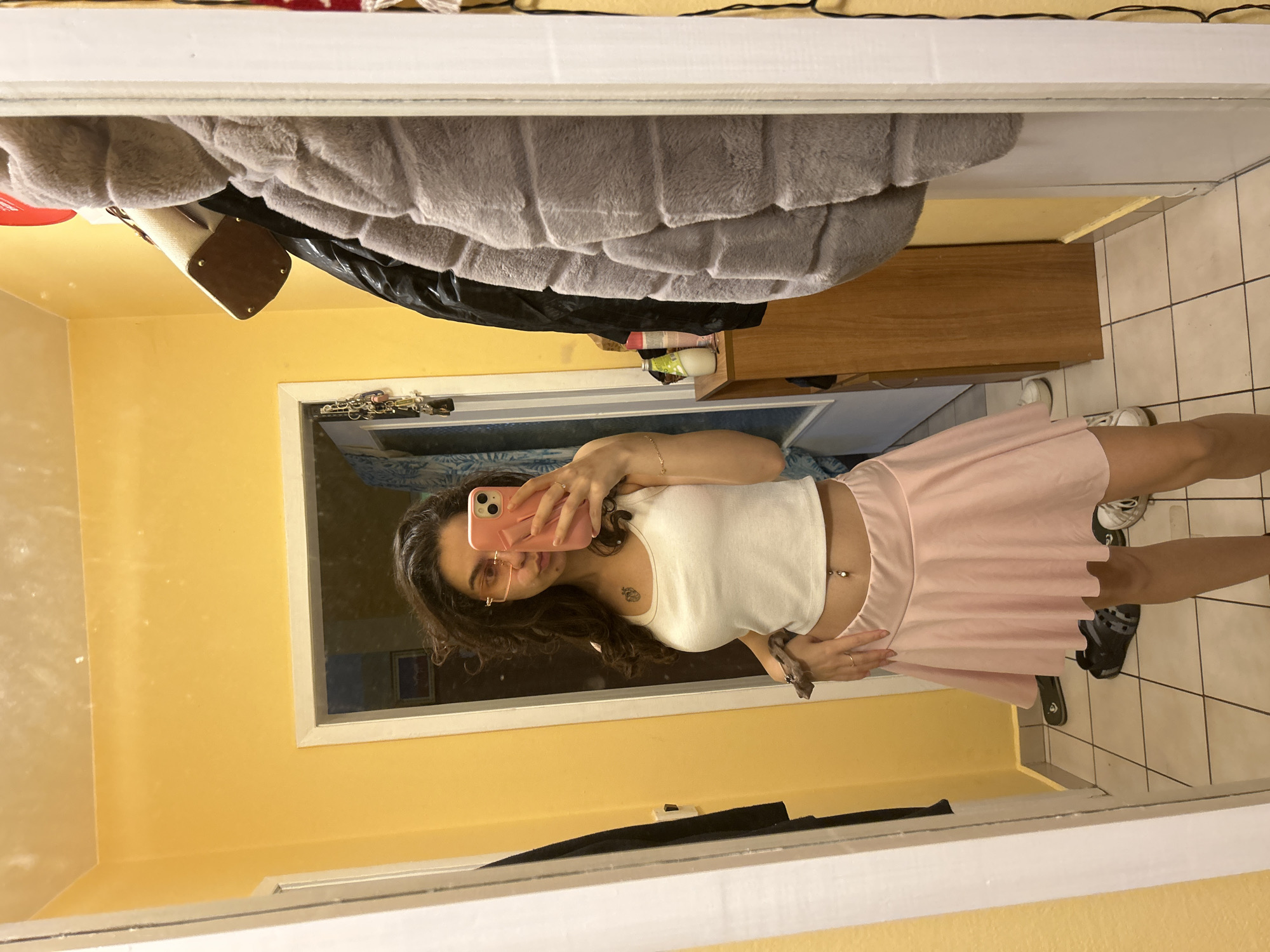 A young woman is taking a selfie in a mirror. She has long, curly brown hair and is wearing glasses, a white crop top, and a light pink skirt. Her face is partially obscured by her pink smartphone. The background appears to be a room with yellow walls and tile flooring. A dresser and some hanging clothes are visible in the upper part of the mirror's reflection. The exact location in Sofia, Bulgaria, cannot be determined from the image.