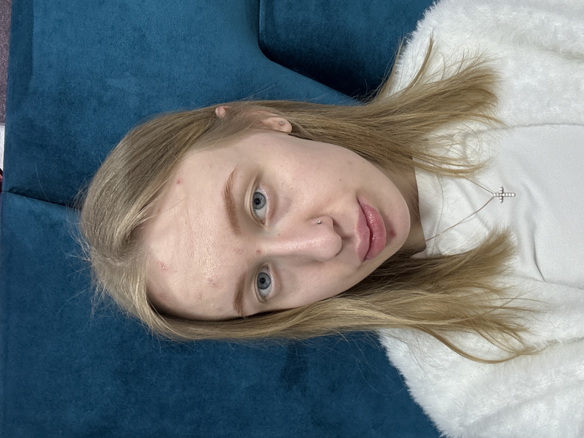 A fair-skinned young woman with blonde hair and blue eyes is lying on a blue surface, possibly a couch. She has a nose piercing on her left nostril and is wearing a delicate silver necklace with a cross pendant. She appears to have some skin blemishes or acne on her forehead. She is wearing a white garment. The location of Rostov-on-Don, Russia, is not discernible from the image.