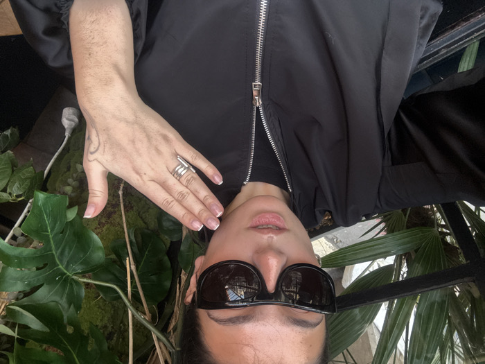 A person wearing black sunglasses and a black jacket is centered, with the image oriented upside down. Their hand, adorned with a silver ring and a tattoo, rests on green plants. The backdrop includes a mix of leafy plants. The individual's face is partially visible, with a focus on the sunglasses and lips. A silver zipper runs down the jacket's front. The surroundings suggest an outdoor or garden setting.