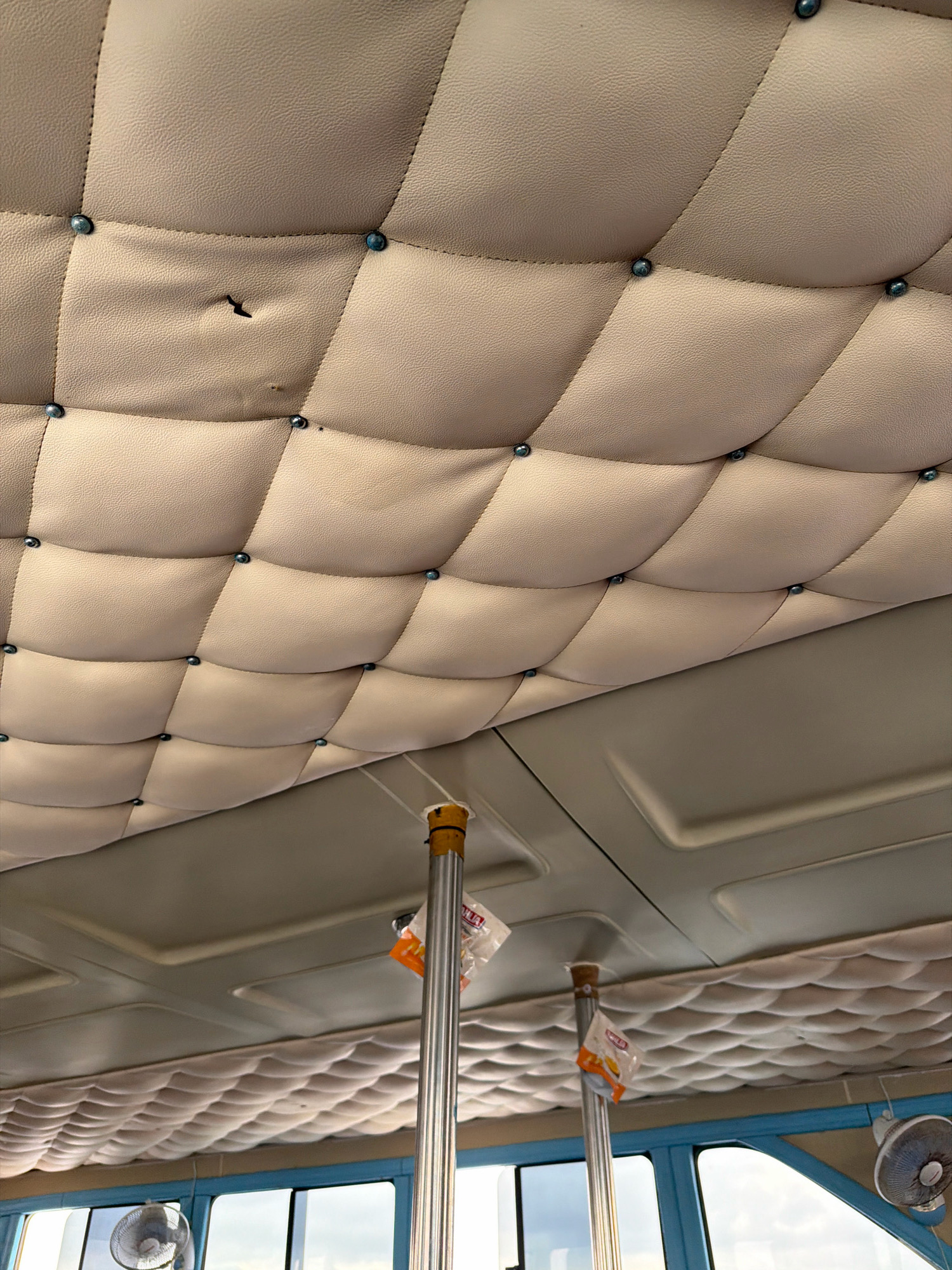 This is a close-up, low-angle shot looking up at the padded ceiling of a vehicle, likely a bus or van. The ceiling is upholstered in a light-colored, textured material, possibly faux leather, with a tufted diamond pattern secured by small, dark blue studs. Several squares of the padding appear slightly puffed out, creating a quilted effect. In the upper left portion of the image, there is a small tear or puncture in the padding, revealing a dark hole.
Below the primary padded ceiling, there is a smooth, lighter gray paneling, also with a subtly embossed pattern. Two vertical, silver poles with yellow and orange elements at their tops extend down from the ceiling, suggesting they are support structures or part of the vehicle's interior. Small, plastic-wrapped items, possibly snacks or amenity kits, are attached to these poles.
In the lower part of the image, the ceiling continues with another section of padded material, this time with a smaller, more rounded pattern of indentations. Through a window frame visible at the bottom, parts of a blue structure and a small, dark electric fan can be seen. The overall impression is of the interior of a vehicle designed for passengers. There is no visible indication of a specific location in the city of Unknown, Indonesia.
