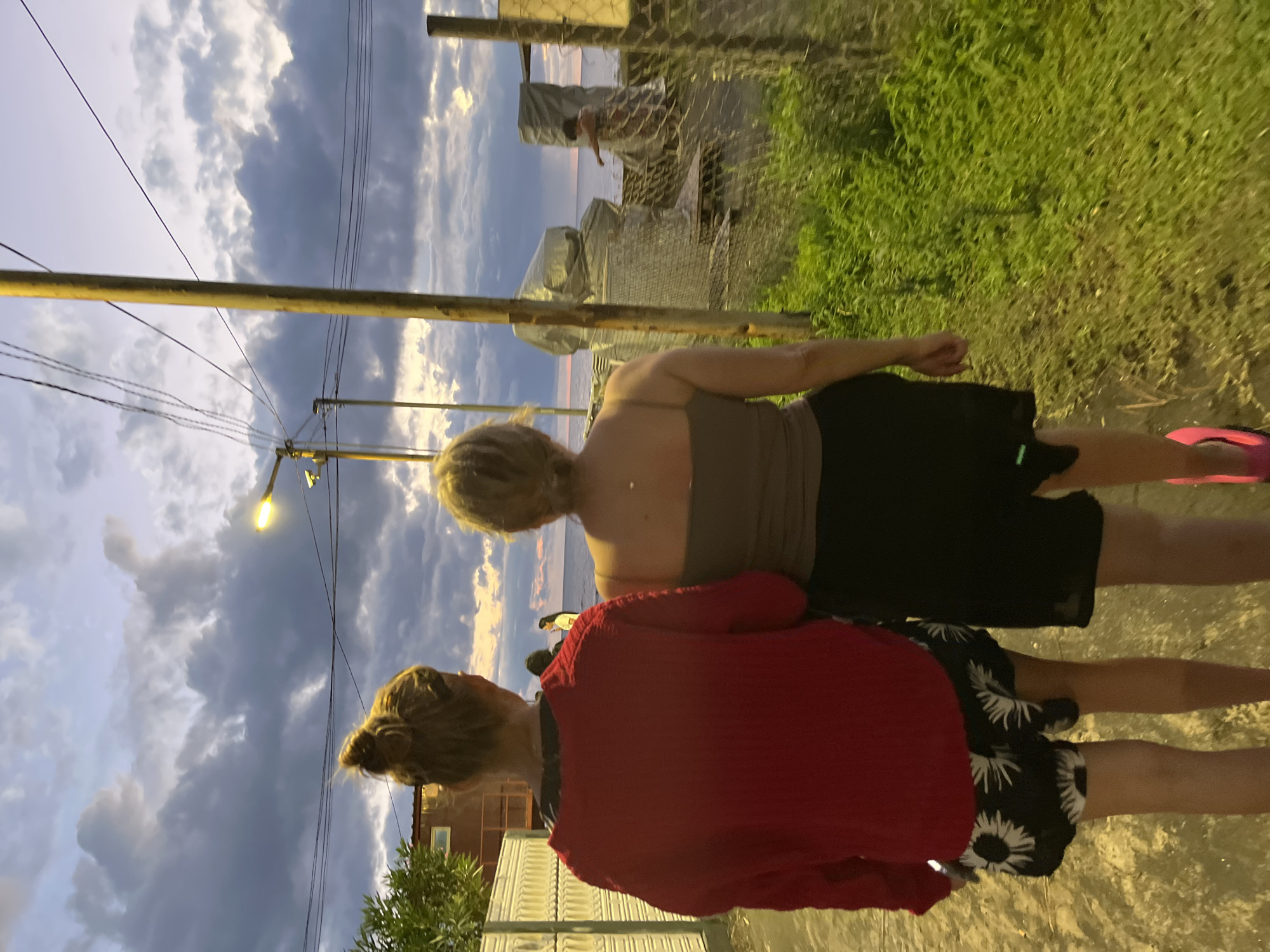 In Ureki, Georgia, two individuals walk away from the camera on a dirt path near the Black Sea. The person on the left wears a red long-sleeved top and black shorts with a white floral pattern. The person on the right wears a grey tank top and black shorts. The sky is cloudy with the sun setting. In the background, a fence encloses an area with construction materials covered in plastic sheeting. Further in the distance, the Black Sea is visible with two other individuals standing near the water. The overall scene depicts a casual evening stroll along the coast.