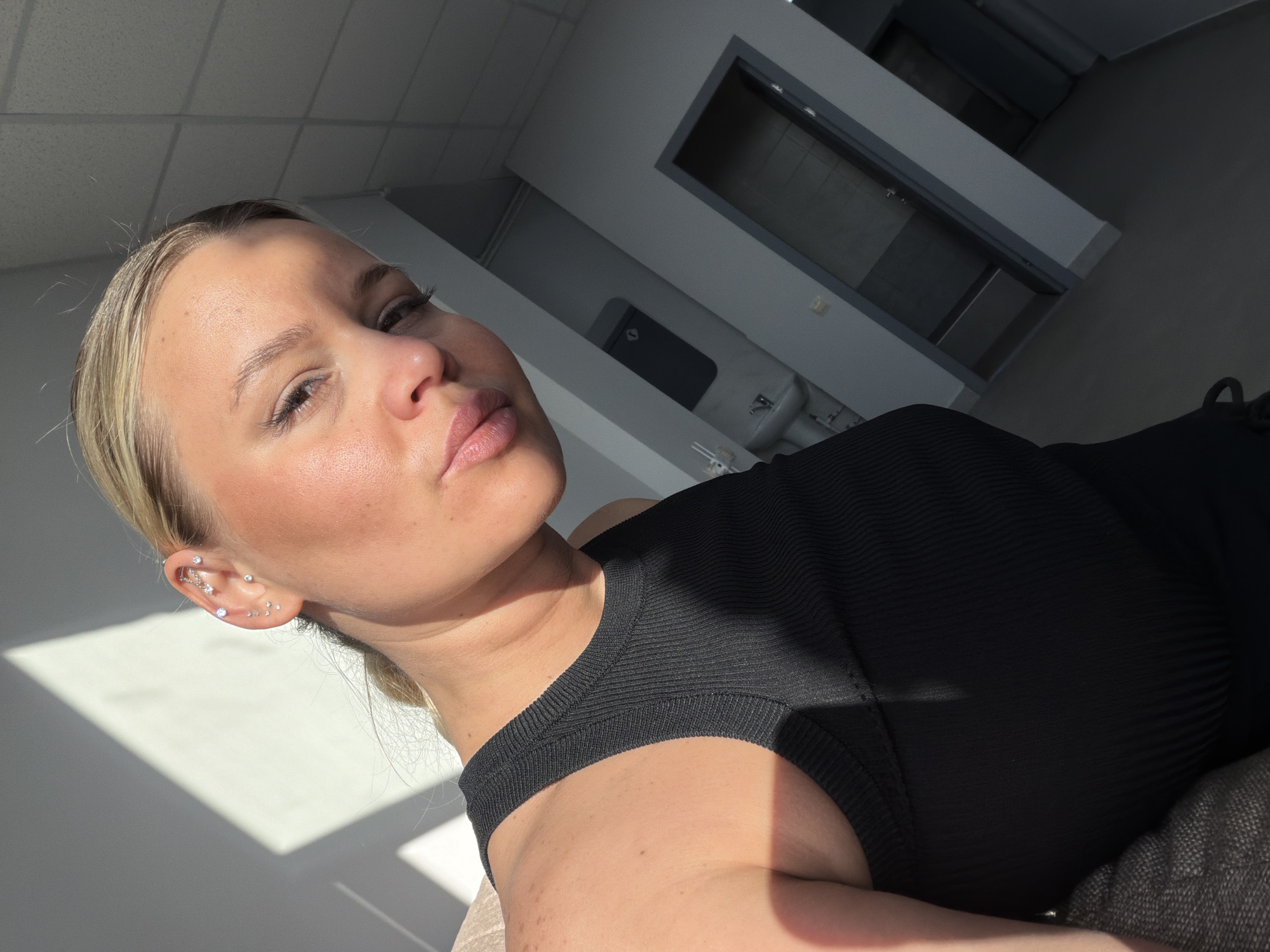 A close-up, eye-level shot of a fair-skinned blonde woman indoors. She is wearing a black, ribbed top with a rounded neckline. Her ear is adorned with multiple piercings. The background includes a section of a gray-painted wall illuminated by sunlight streaming in from a window on the left. The ceiling appears to be white and has a tiled or paneled texture. In the upper-right portion of the photo, there are gray-framed doors. The scene does not contain enough information to determine the location in the city of Sarapul, Russia.
