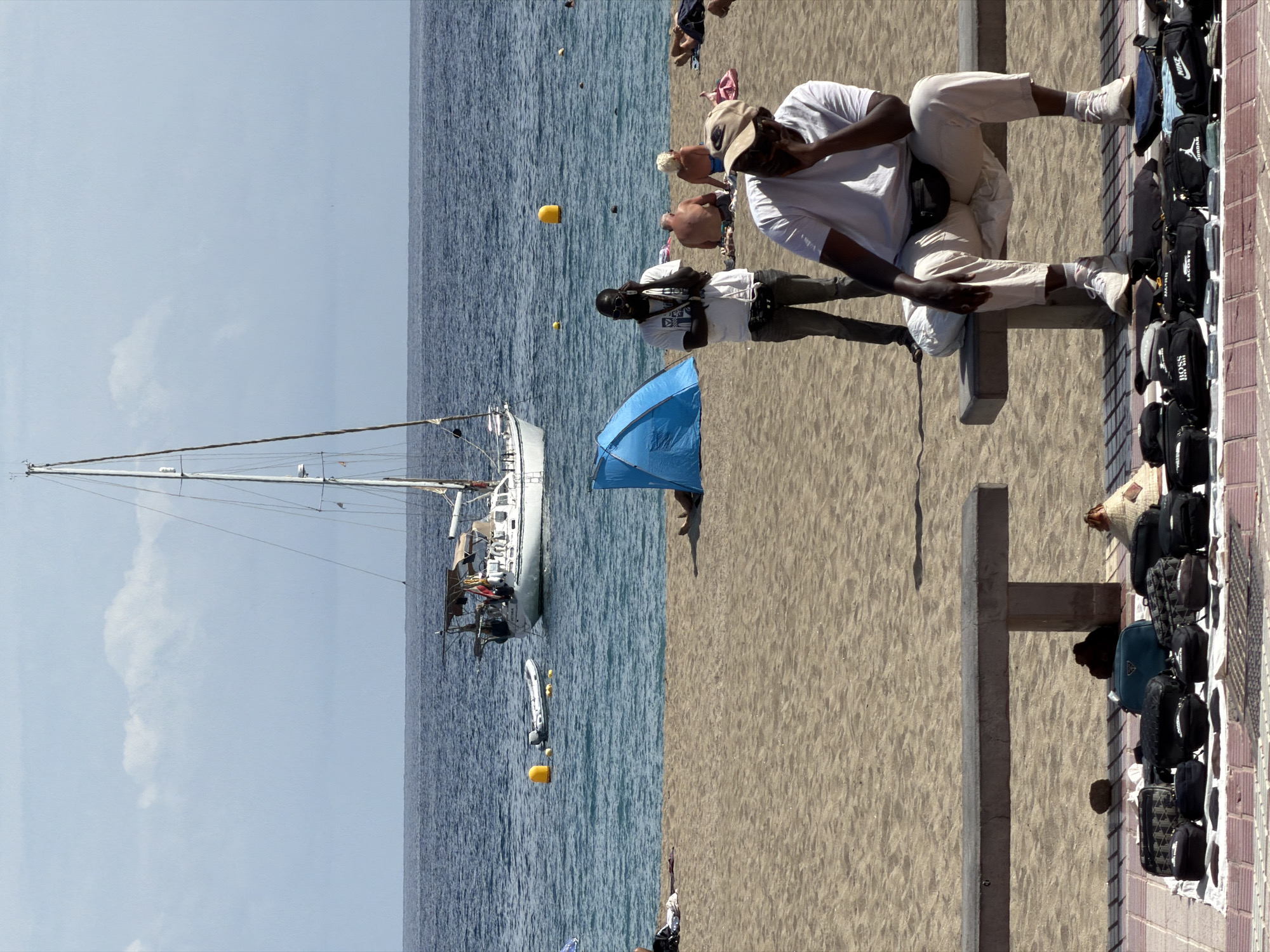 This image captures a vibrant beach scene in Los Cristianos, Spain, on a bright, sunny day. The clear blue sky is visible above a tranquil, blue-green ocean with small waves. A stretch of light brown sand occupies the foreground and mid-ground.
Several people are present, engaging in various activities. In the mid-ground, a man wearing a white t-shirt, light pants, and a baseball cap is seated on a low, concrete barrier or bench, possibly observing the scene or using a device. To his left, another man in a white t-shirt, dark pants, and sunglasses stands with his arms crossed, facing towards the water. A blue beach umbrella is partially visible behind him on the sand. Further in the background, other individuals are scattered across the beach and in the water, enjoying the sun and sea. One person near the water wears a blue swimsuit.
In the ocean, a large white sailboat with a tall mast is anchored on the left side of the frame, with a small dinghy or inflatable boat nearby. Two yellow buoys float in the water closer to the shore.
On the far right, alongside a paved walkway and a low brick wall, a display of merchandise is set up. Numerous bags, predominantly dark-colored fanny packs and larger bags, are laid out on white mats. Visible brand names on these items include "NIKE," "PUMA," "LACOSTE," and "BOSS." A vendor, partially obscured, appears to be attending to the display.
The time of day is likely midday, indicated by the high sun and distinct shadows. The overall atmosphere suggests a lively and typical beach day with both leisure and commercial activities taking place.
