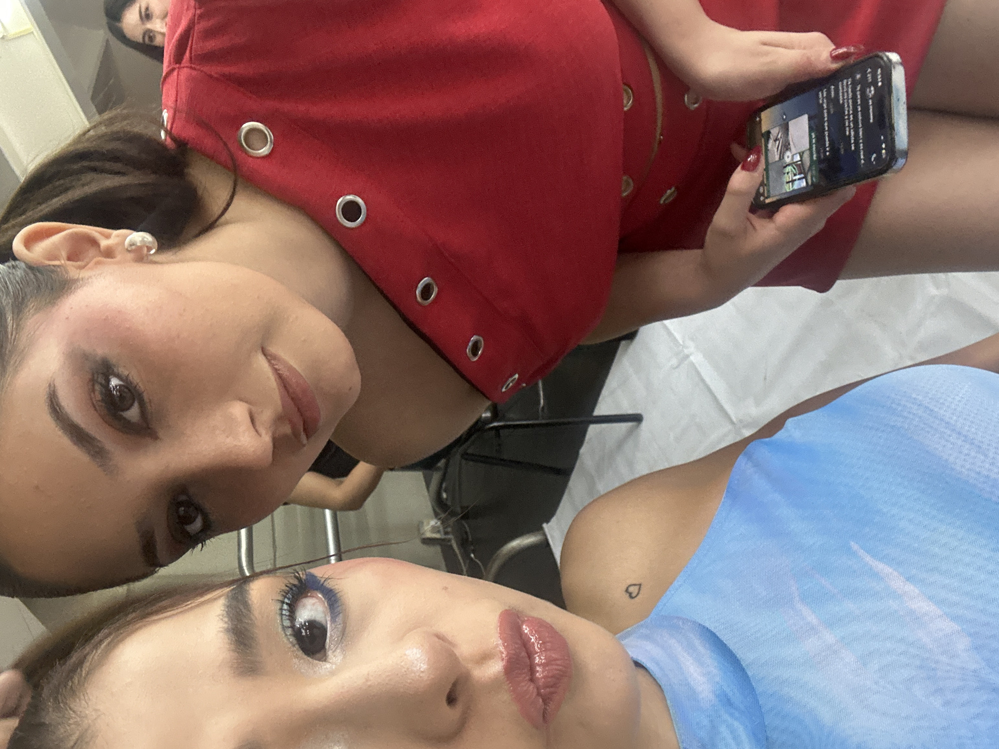 This close-up, overhead shot captures two women in an indoor setting, likely in San Bernardino Tlaxcalancingo, Mexico. The scene appears to be a salon, spa, or backstage area.
In the upper left, one woman leans forward, gazing directly into the camera with a subtle smile. She has dark hair and wears a red garment distinguished by numerous silver grommets. A silver stud earring is visible on her left ear. Her hand, with red-painted nails, holds a smartphone displaying what appears to be a social media feed or image gallery with small thumbnails and unreadable text. A third person is partially visible behind her head.
The second woman occupies the lower right of the frame, lying down and looking upwards. She wears a light blue, possibly disposable, top, revealing a small, black heart tattoo on her left shoulder. Her face is made up with striking blue eyeshadow and glossy, nude-pink lips, giving her a neutral expression.
The background consists of a white, textured surface, resembling a sheet or paper, and dark, indistinct furniture. The intimate framing suggests an activity involving close interaction, potentially a beauty treatment, photo session, or casual moment among friends.