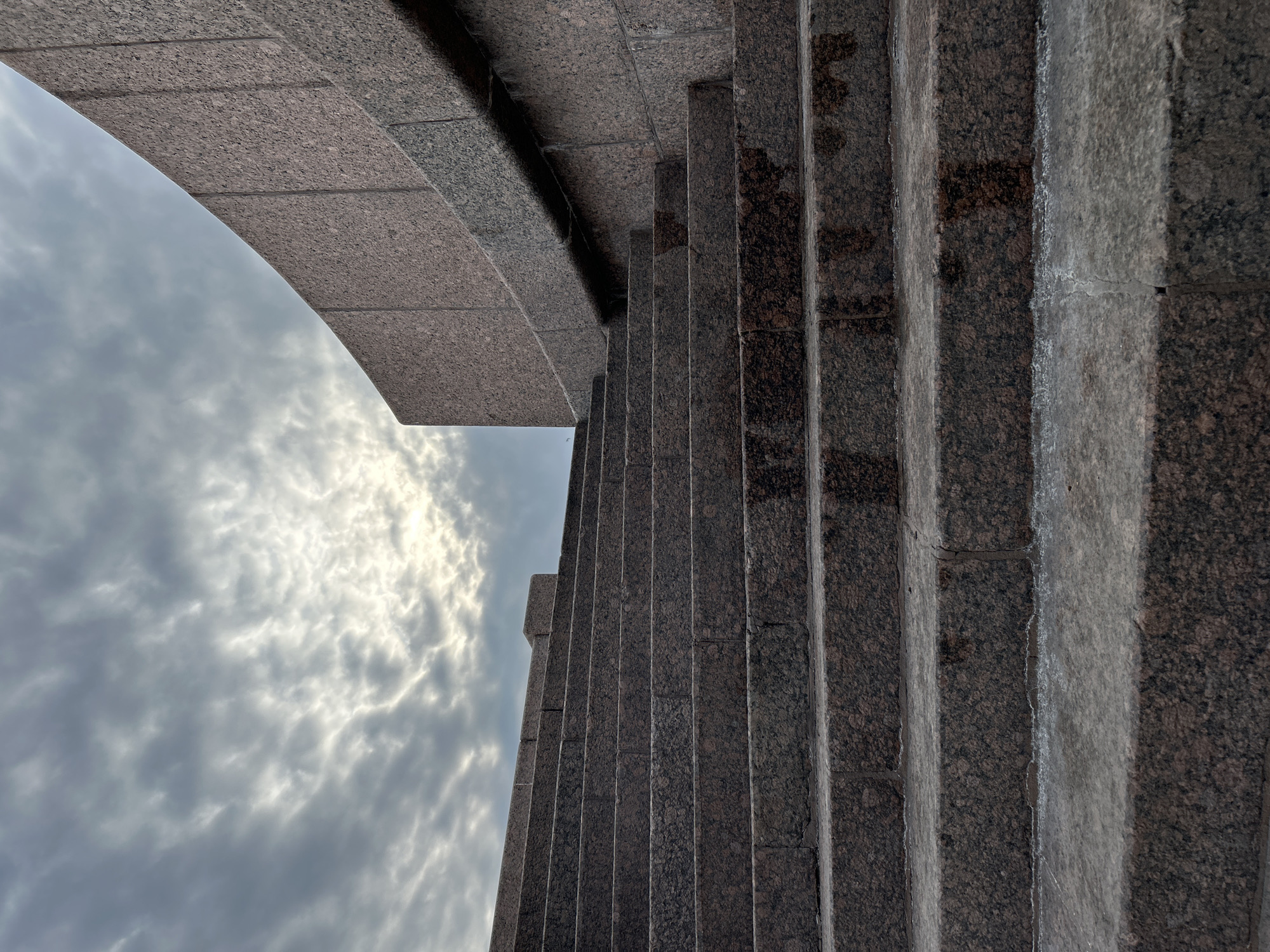 The image depicts a low-angle view of a granite structure, likely part of an architectural element in Saint Petersburg, Russia. The structure features a curved arch on the left side of the image with linear columns on the right, all constructed from a dark, speckled granite. The background consists of a cloudy sky with patches of light breaking through. There is no presence of people, subjects, objects, actions, or interactions, focusing solely on the architectural and environmental aspects.