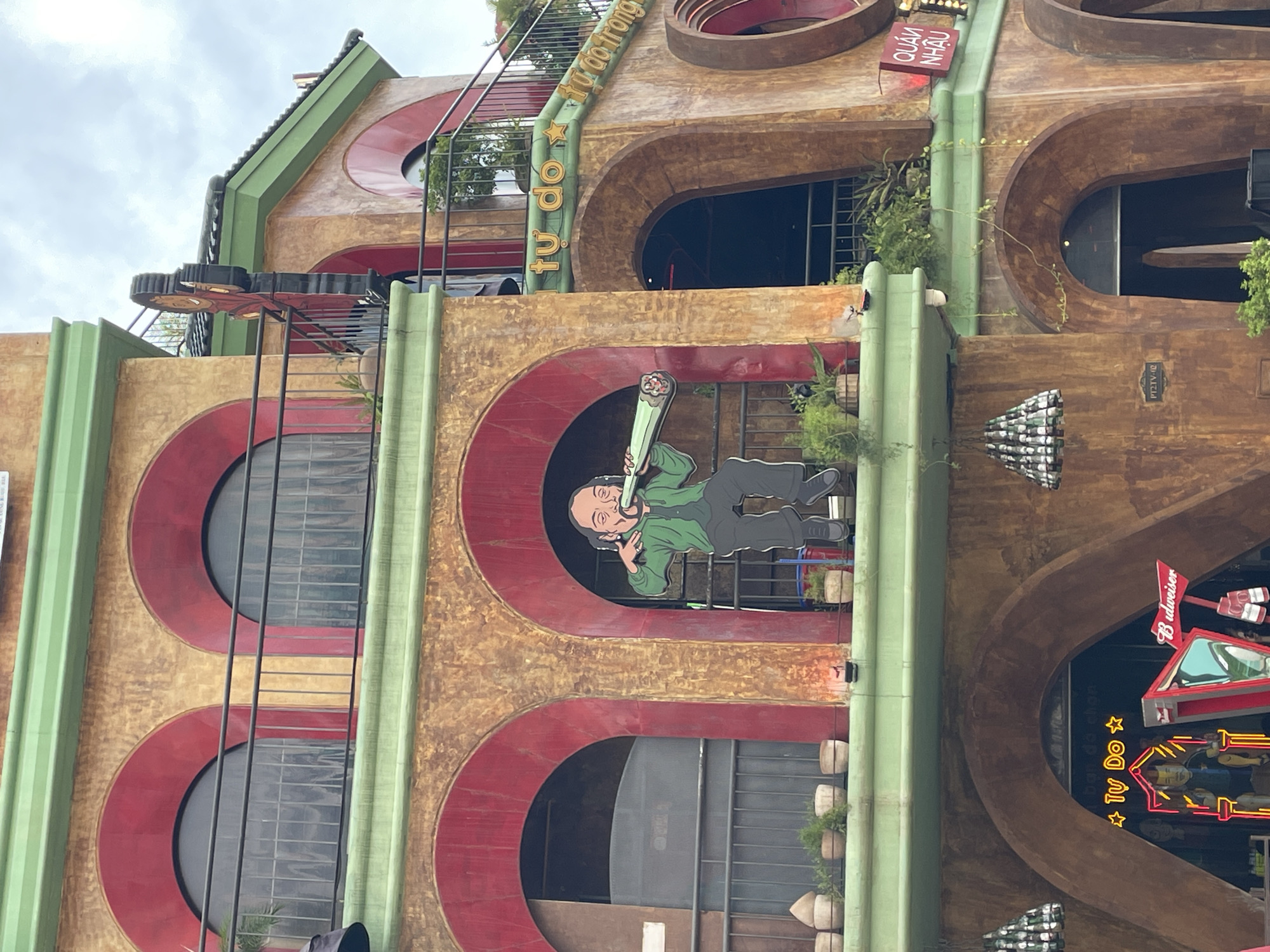 The image shows the exterior of a multi-story building, likely a restaurant or bar, with an earthy-toned facade featuring red arched windows and balconies. A prominent cutout figure of a person smoking what appears to be a large cigarette is positioned in one of the arched openings. Green trim runs along the edges of the building's levels.
On the upper right side of the building, text in yellow lettering is visible, which reads "TU DO" and "tu do trong". A red sign with white lettering that reads "QUÁN NHẬU" is also present.
Below, at the ground level, an arched entrance reveals neon signage. One sign spells out "TU DO" with stars on either side, and another sign is a Budweiser logo. The building is decorated with hanging light fixtures made of bottles. The overall setting appears to be a street in Nghĩa Trụ Commune, Vietnam, judging by the presence of Vietnamese text on the signage and the architectural style. The sky is overcast.