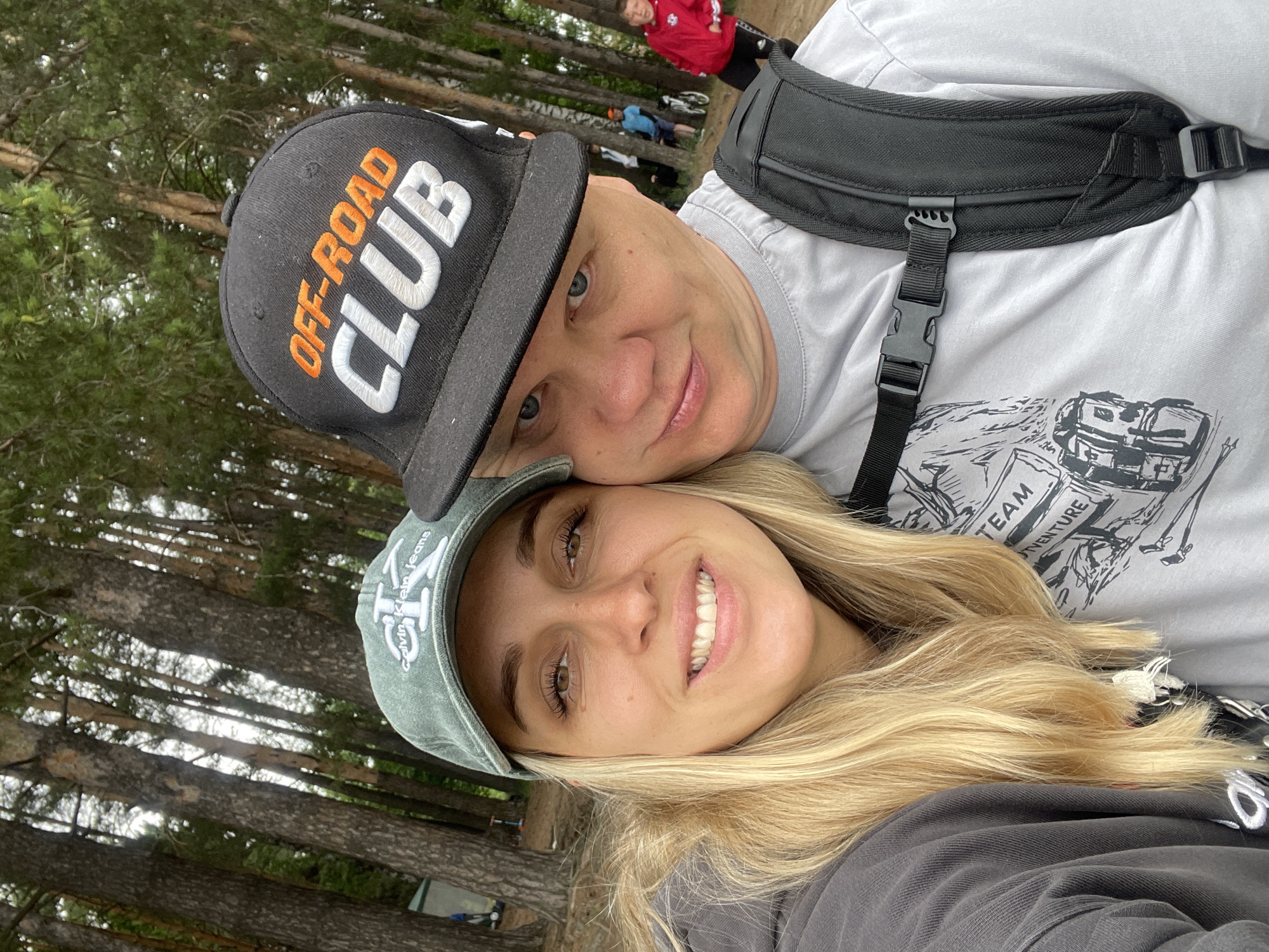A selfie of a couple taken outdoors in a forest setting. The woman on the left is wearing a green baseball cap with "Calvin Klein Jeans" embroidered on it and a grey hooded sweatshirt. She has blonde hair and is smiling. The man on the right is wearing a black baseball cap with "OFF-ROAD CLUB" embroidered on it and a grey t-shirt with a backpack. He has blue eyes and is looking towards the camera. In the background, there are tall pine trees and other people can be seen, including a boy in a red jacket. The location is the city of Chaikovsky, Russia.
