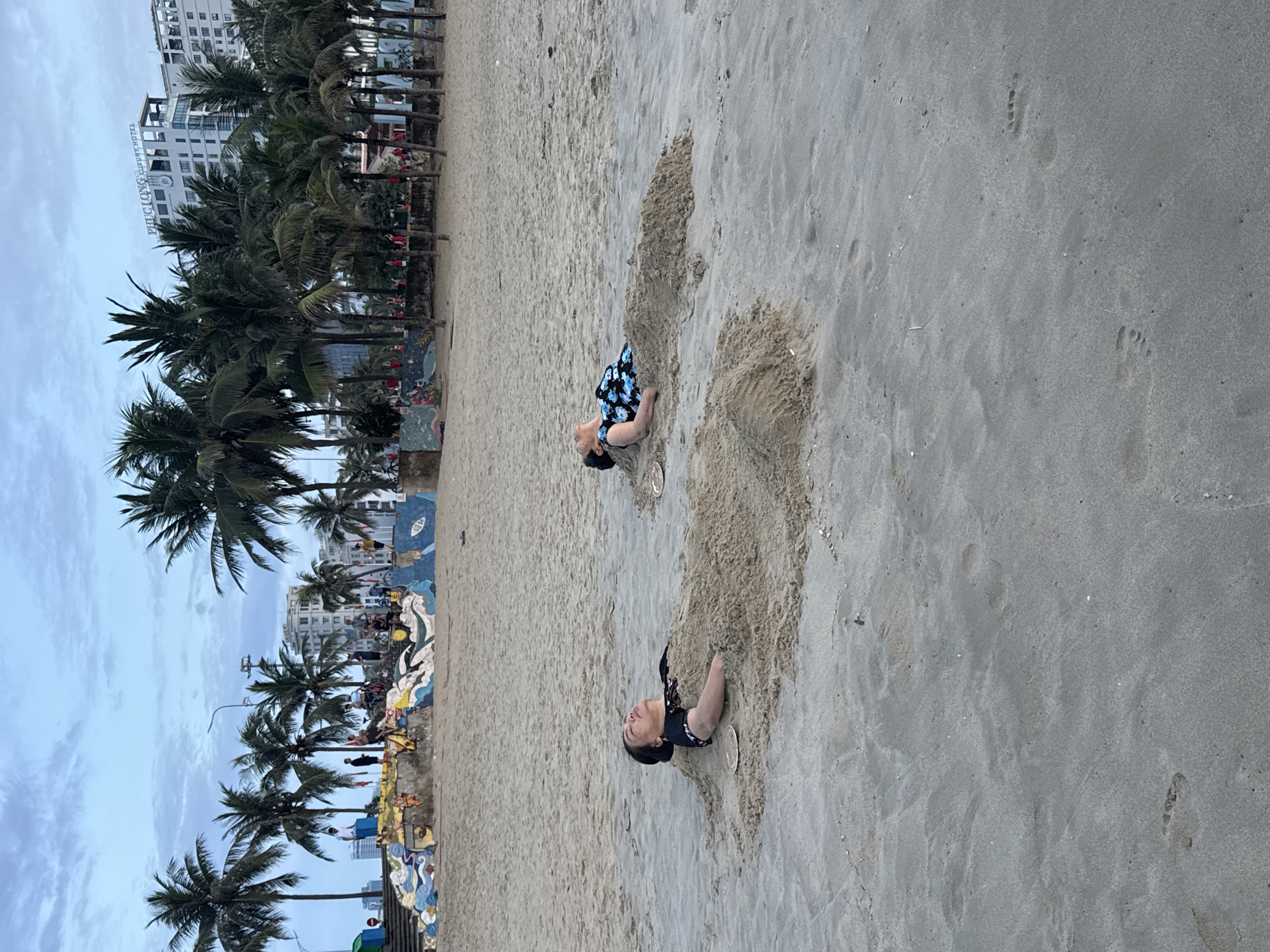 Two people are buried in the sand on a beach. The people are buried up to their necks. In the background, there are palm trees, a building, and a mural. It is in Đà Nẵng, Vietnam.