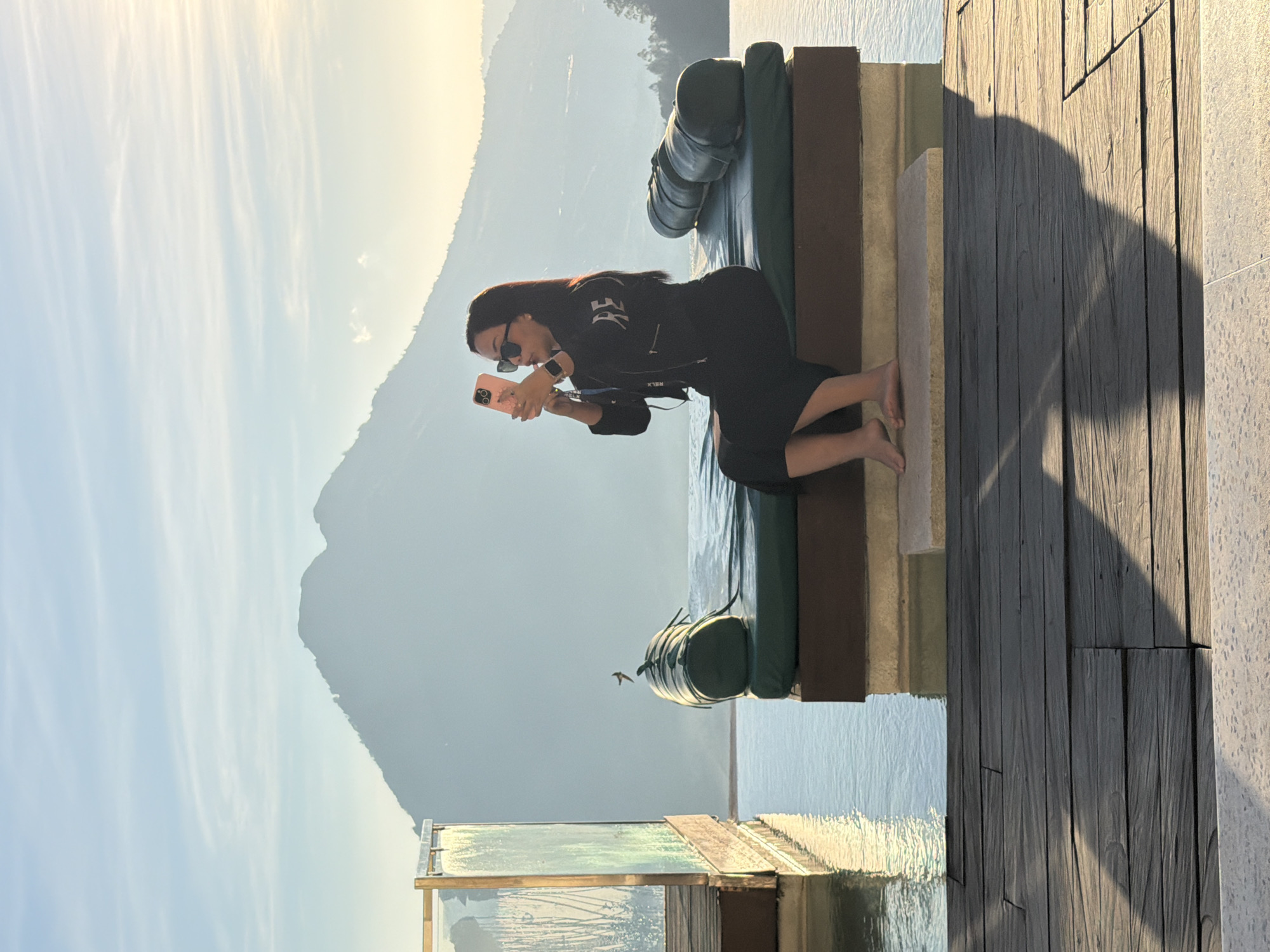 This image captures an outdoor scene at a resort in Kintamani, Indonesia, overlooking a mountainous landscape, likely Mount Batur. A single female individual, with dark hair, sunglasses, and dressed in a black long-sleeved top with "RELAX" written in white and a black midi-skirt, is seated barefoot on a dark green padded daybed. The daybed has a dark brown base and is positioned at the edge of an infinity pool. She is holding a pink smartphone up to take a picture, facing slightly to the right. To her left, a large, cylindrical, rolled-up bolster cushion rests on the daybed. Above this cushion, a small, dark bird is captured in flight. To the far left, a glass partition with a golden frame, featuring a patterned design, extends into the pool. The infinity pool's calm surface reflects the sky and mountains. In the background, multiple layers of hazy, blue-grey mountains rise, with a prominent, large peak dominating the central view. The sky above is light blue with delicate horizontal cloud formations, illuminated by golden light, indicating either sunrise or sunset. The foreground consists of a dark wooden deck with prominent shadows cast upon it.