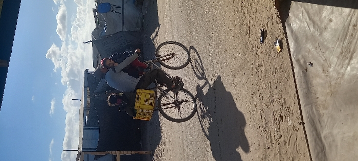 A daytime scene in Khan Yunis, Palestinian Territory, depicts an adult male cycling a bicycle with two children on a dirt and sand path. The adult, wearing a light-colored shirt and dark pants, is positioned over the bicycle frame. A child, identifiable by a pink head covering, sits in front of the adult, while a second child, dressed in dark attire, occupies a yellow multi-compartment plastic crate secured to the bicycle's rear rack. Strong shadows from the bicycle and its occupants are cast to the left on the sunlit ground. The background consists of multiple makeshift shelters and tents constructed from various materials, including tarpaulins and fabric, under a blue sky with scattered white clouds. Small pieces of debris are visible on the ground in the foreground.