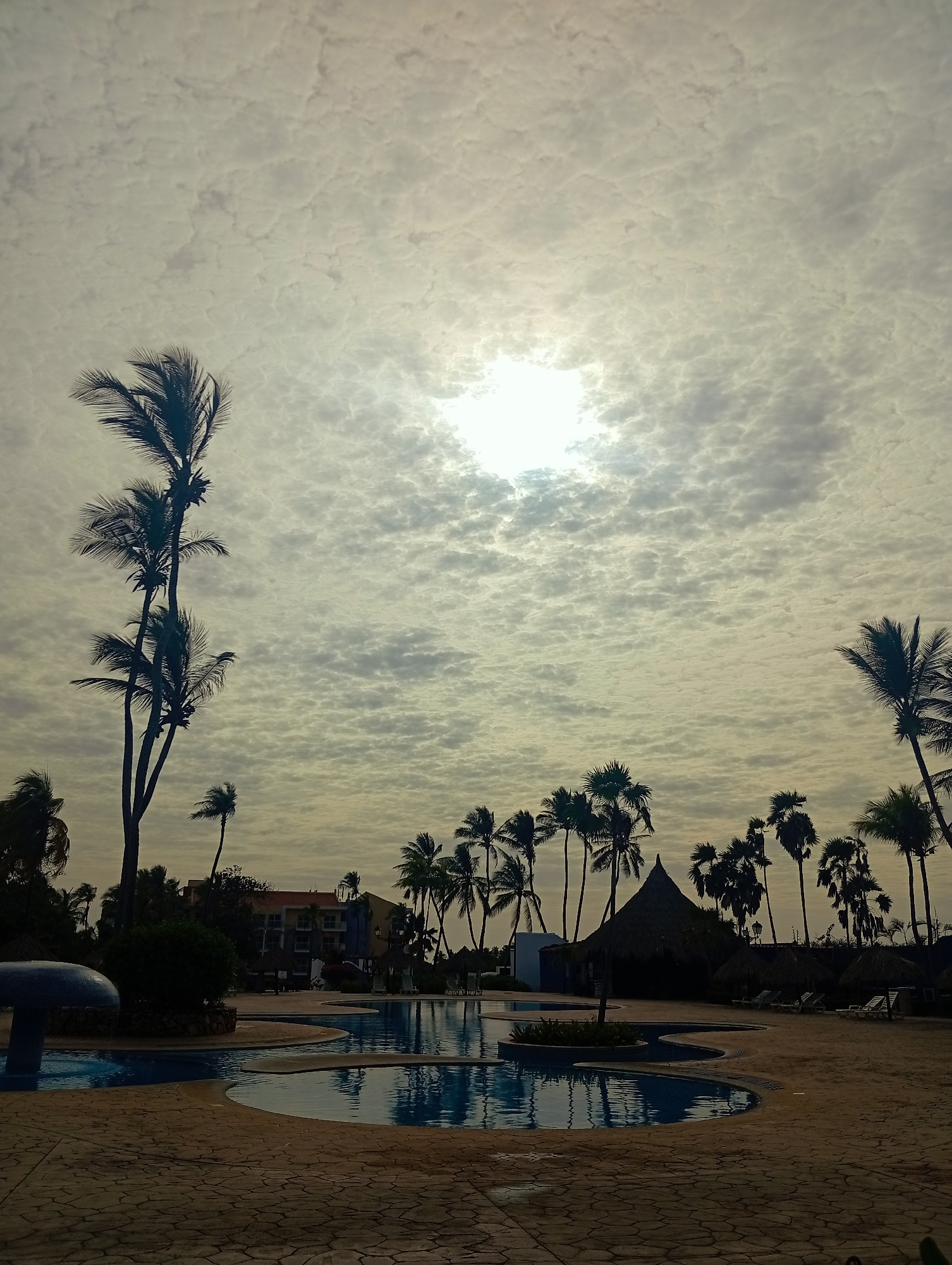 The image shows a swimming pool area in Playa El Agua, Venezuela, at dusk or dawn. The sky is overcast with a thin layer of clouds, obscuring the sun. Several palm trees line the area around the pool. A building with multiple floors is visible in the background. A thatched-roof structure stands near the pool, along with several lounge chairs. The pool water reflects the cloudy sky. The foreground shows the paved area around the pool, constructed with stone bricks. A mushroom-shaped structure is partially visible at the edge of the pool. There are no people visible in the image.