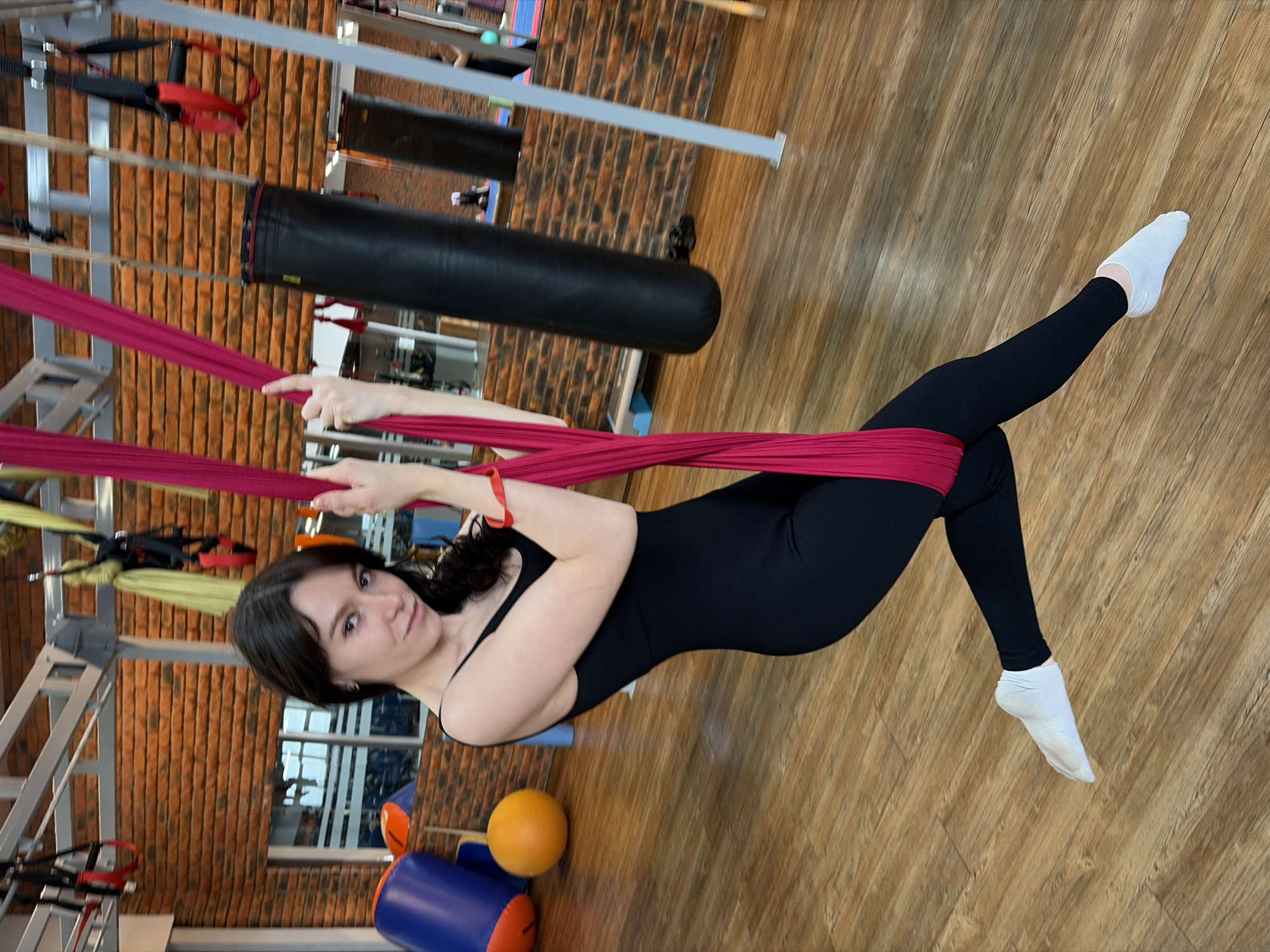 A woman wearing a black leotard and white socks is suspended from a red aerial yoga hammock in a gym. She is holding onto the hammock with both hands and has one leg extended straight out in front of her. The gym has wood flooring and a brick wall. A black punching bag is visible in the background. Based on the presence of Cyrillic text and the reported location, the scene is likely in Chaikovsky, Russia.