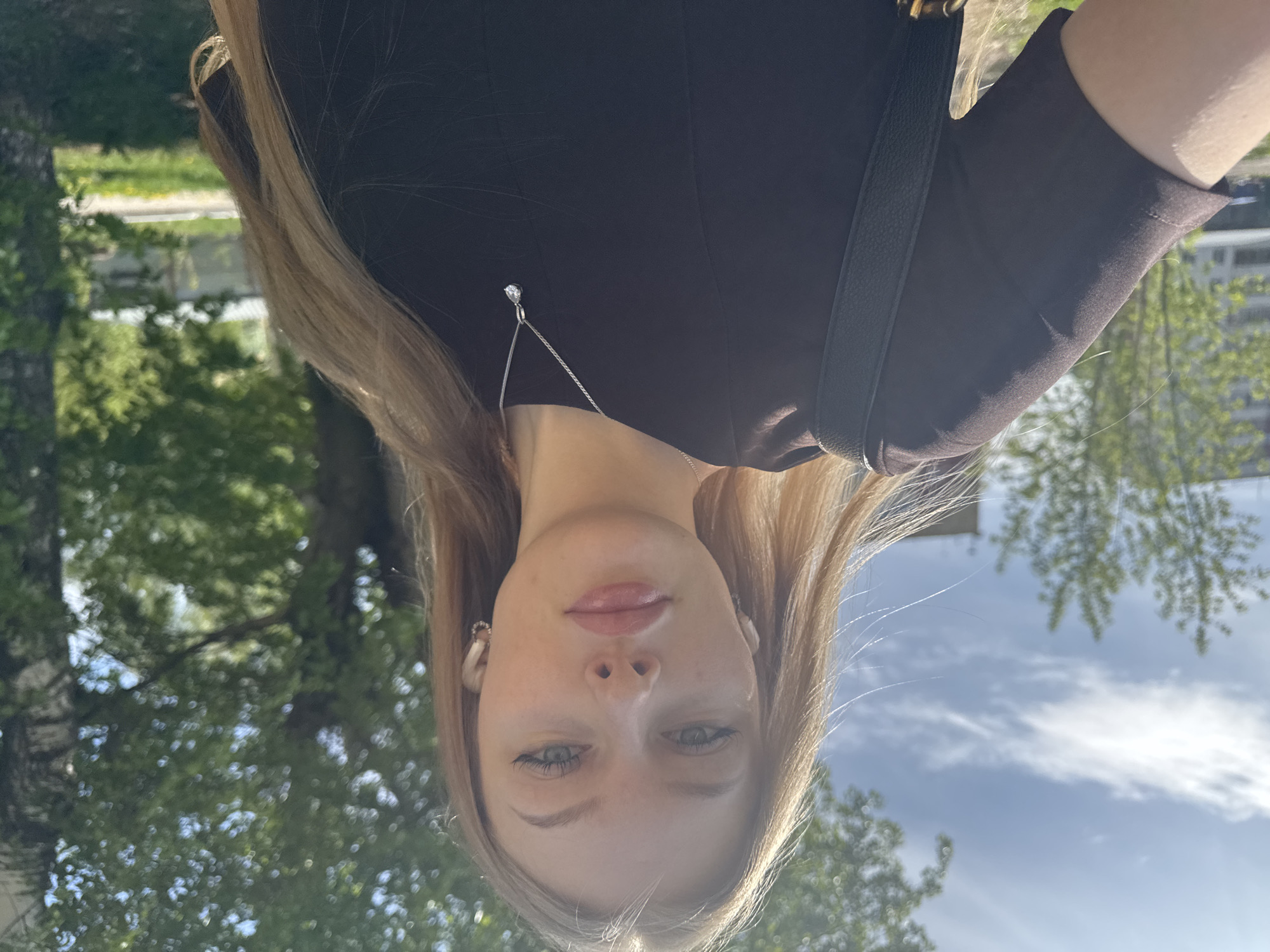 A young, fair-skinned woman with long blonde hair, light-colored eyes, and pink lips is the primary subject of this outdoor selfie. She is wearing a brown top and a silver necklace with a small pendant. A black bag strap is visible over her shoulder. The background consists of green trees and a partially visible blue sky with clouds. Based on visual cues like architecture and vegetation, the location appears to be Perm, Russia. There are buildings visible behind the trees. The image is oriented upside down.