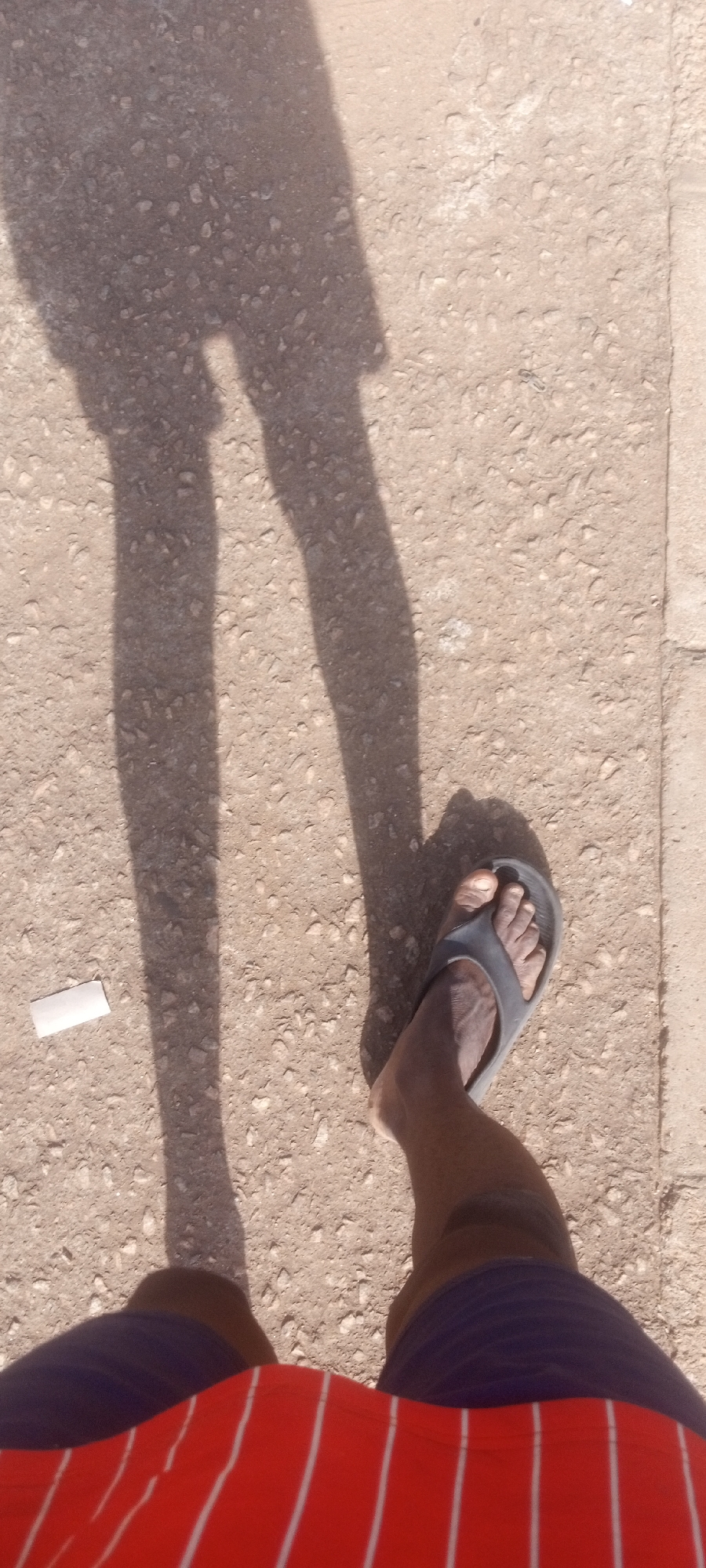 A first-person view looking down at a person standing on a concrete or paved surface, presumably in Sabon Tasha, Nigeria. The person is wearing shorts; the visible portion is red with white vertical stripes over a navy-blue underlayer. The person is also wearing a dark-grey/black sandal on their right foot. The person's shadow falls onto the ground, indicating bright sunlight. A small, white rectangular object sits near the left leg of the shadow. The paved surface appears rough, with small pebbles or stones embedded in it.