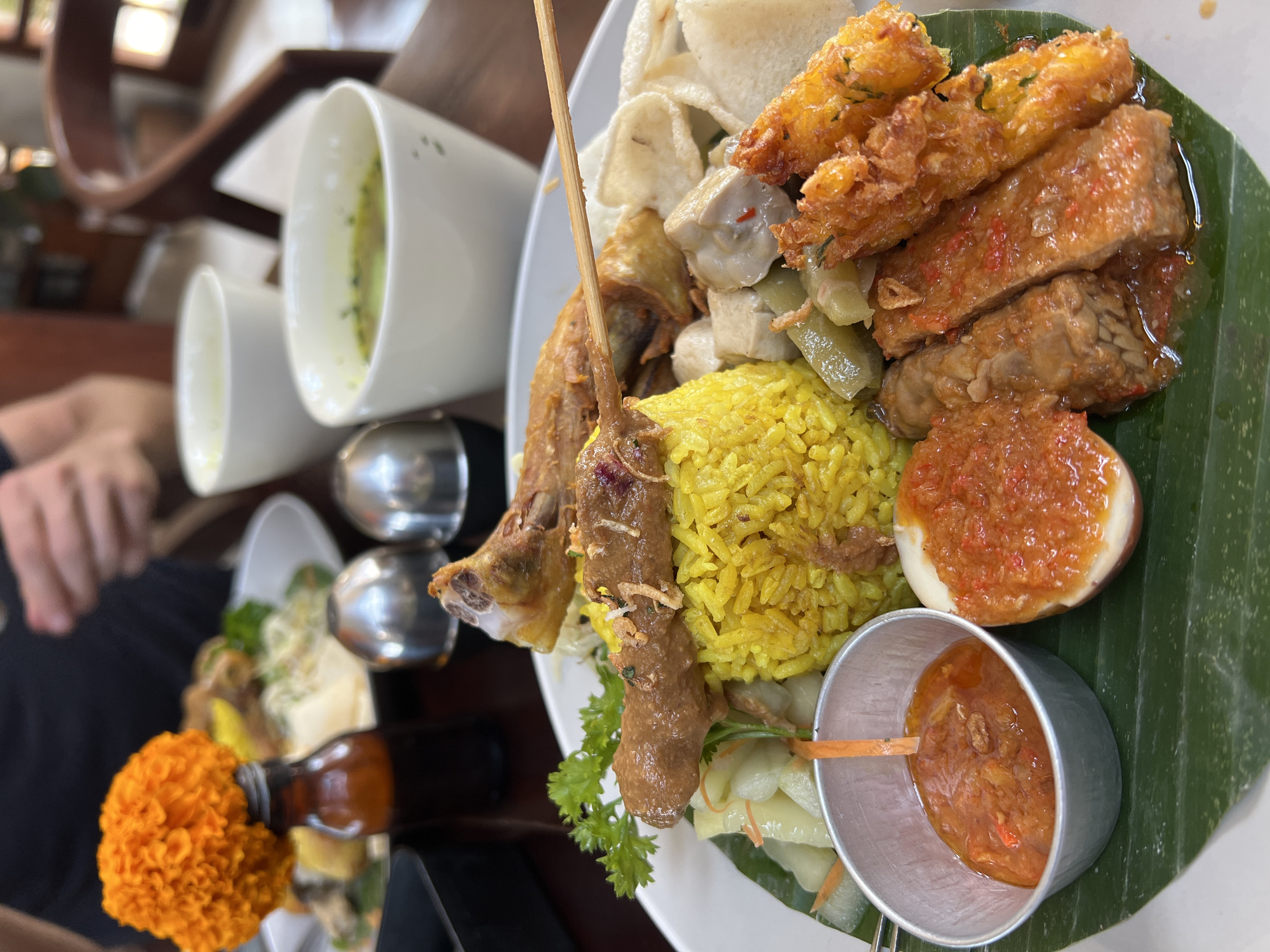 A close-up, eye-level shot shows a platter of Indonesian food, possibly from a restaurant in Ubud District, Indonesia. The platter, placed on a wooden table, features a mound of yellow rice, a skewered meatball with sauce, a piece of fried chicken, a hard-boiled egg cut in half with chili sauce, fried tempeh in chili sauce, fried fritters, crackers, pickled vegetables, and a small metal cup of chili sauce. Two white ceramic bowls of soup are visible in the background, along with two metal shaker lids. An orange marigold flower sits atop a small brown bottle to the left of the platter. In the background, out of focus, are parts of two individuals, one with their hand near their mouth, and a wooden chair.