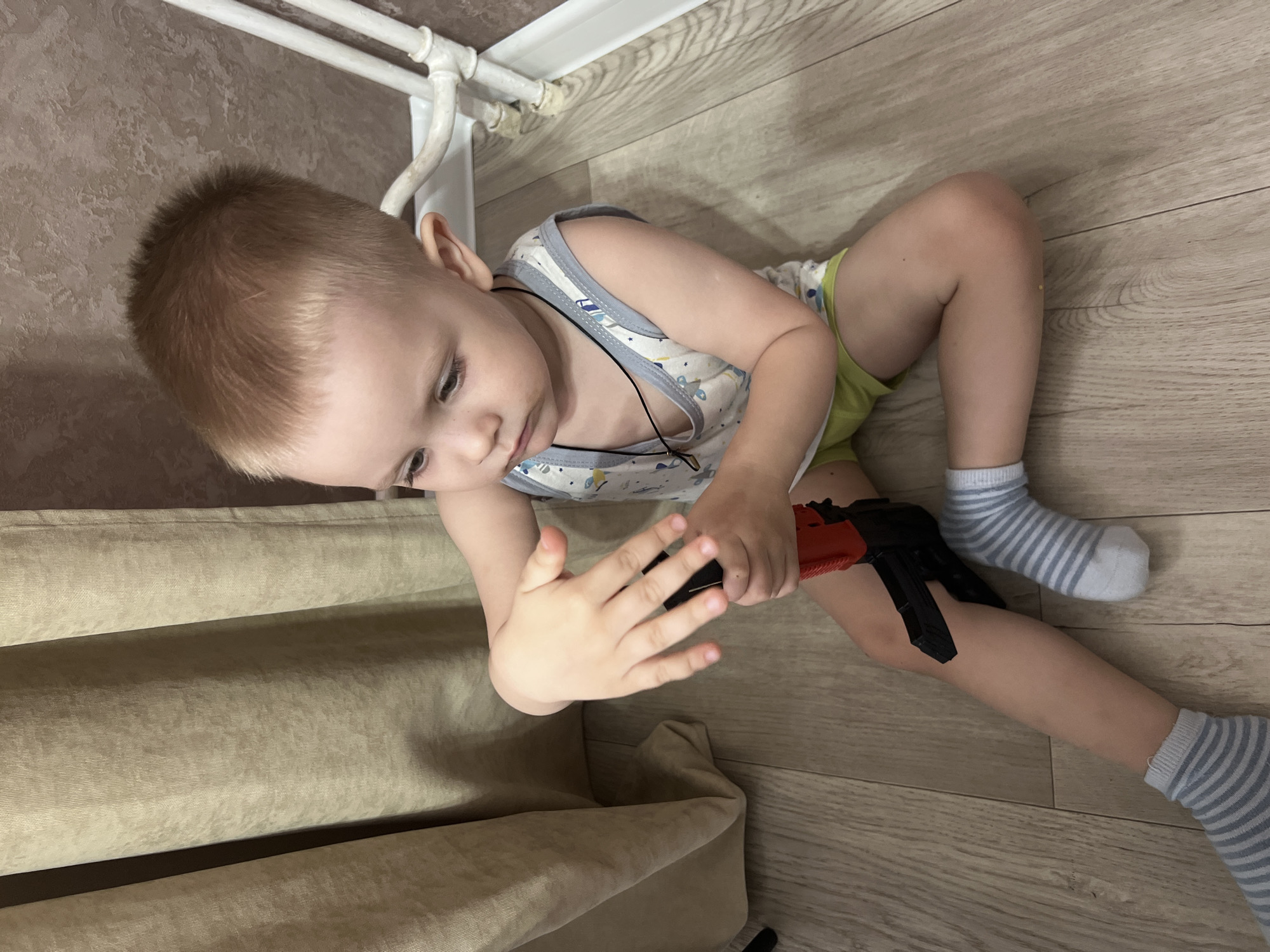 A young child, approximately 2-3 years old, is lying on a light-colored, wood-patterned floor in a room. The child is wearing a light-colored tank top with a blue and white pattern, pale green shorts, and gray and white striped socks. The child has light blonde hair and is holding a black and red toy gun in their right hand, looking down at it. The child's left hand is open with fingers extended. A beige curtain is partially visible on the left side of the image. White pipes are seen along the top edge of the image. The location is Марковский, Russia.