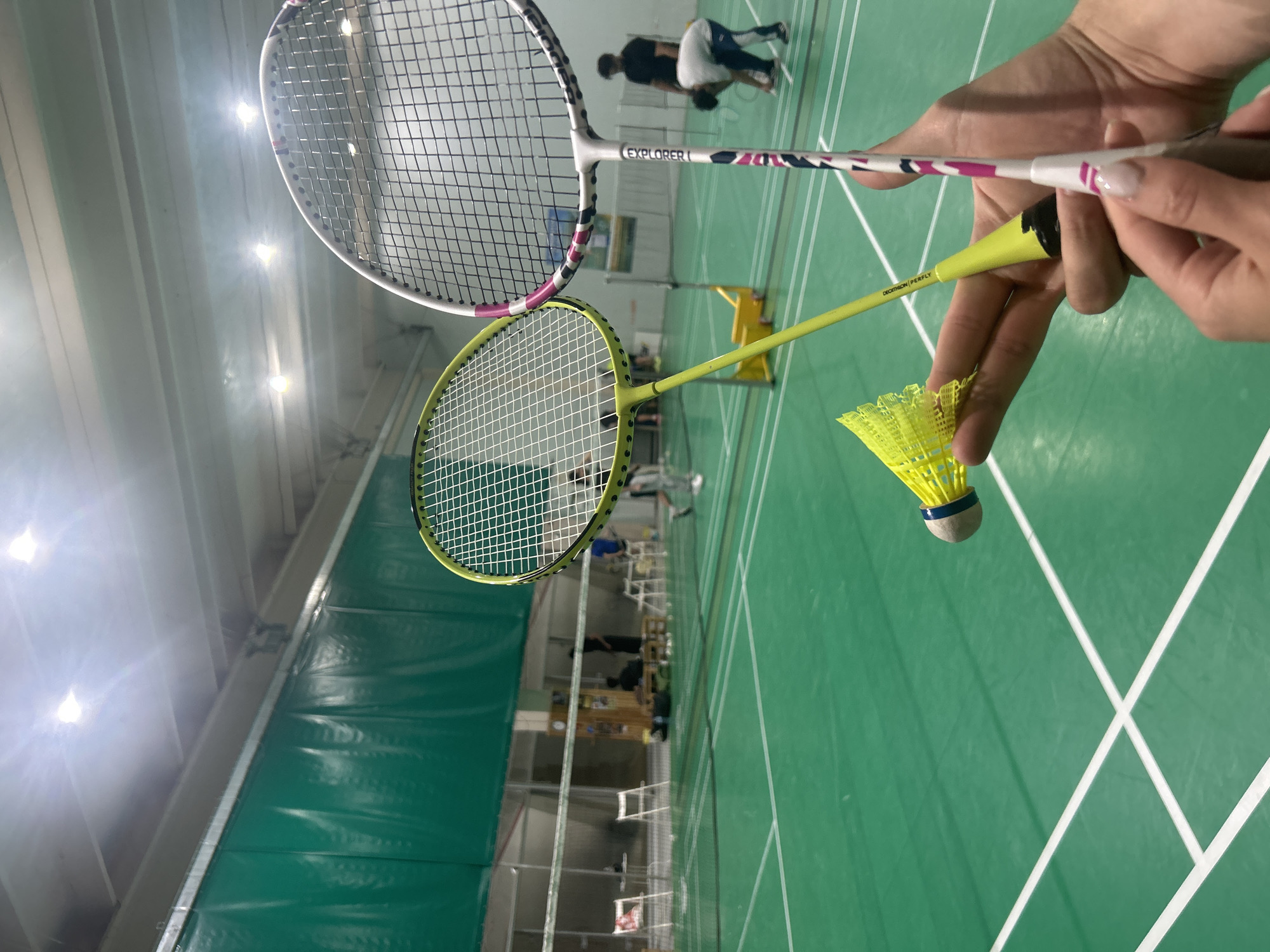 Indoor badminton court, two rackets and a shuttlecock. Sofia, Bulgaria. Two people are playing in the background. One racket is white and pink, brand Explorer. The other racket is yellow, brand Decathlon Perfly. The shuttlecock is yellow. A hand is holding the rackets and the shuttlecock.