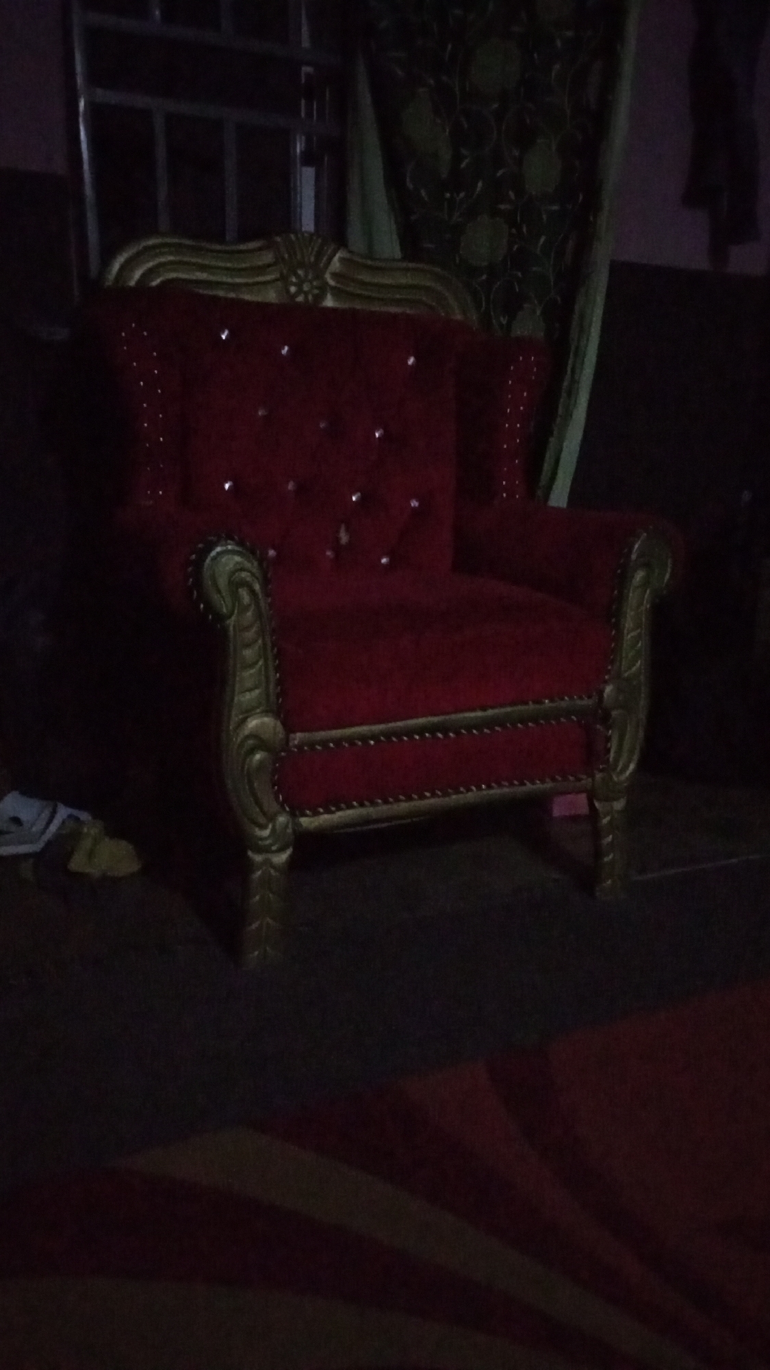 A red, ornate armchair with gold trim is the prominent subject in the scene. The armchair features a high back with button tufting and is positioned on a dark, indistinct surface. The background includes a window with bars, a patterned curtain, and a silhouette of a person. The location is suggested to be Sabon Tasha, Nigeria.
