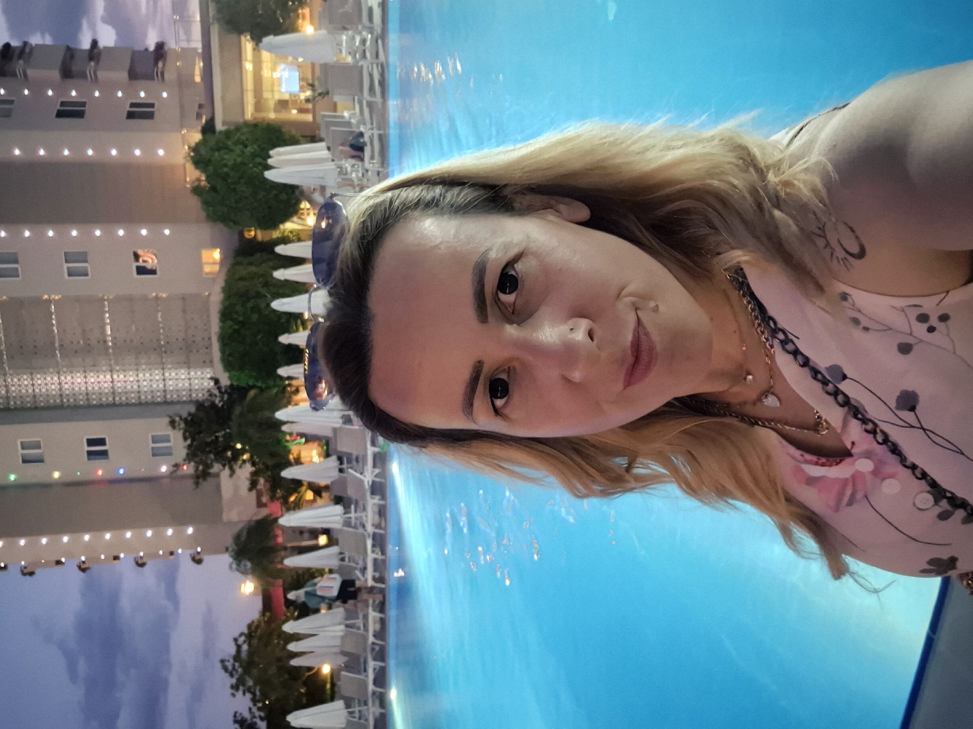 A woman is taking a selfie by a swimming pool at dusk. She is positioned in the foreground, looking directly at the camera with a slight smile. She has blonde hair and is wearing a light-colored, floral-patterned top and a necklace. Behind her, a large, brightly lit hotel building stands tall against a dusky sky. Rows of white lounge chairs and umbrellas are arranged by the vibrant blue swimming pool. Several people are visible in the background, some near the pool and others walking near the hotel entrance. The overall scene suggests a vacation or resort setting.