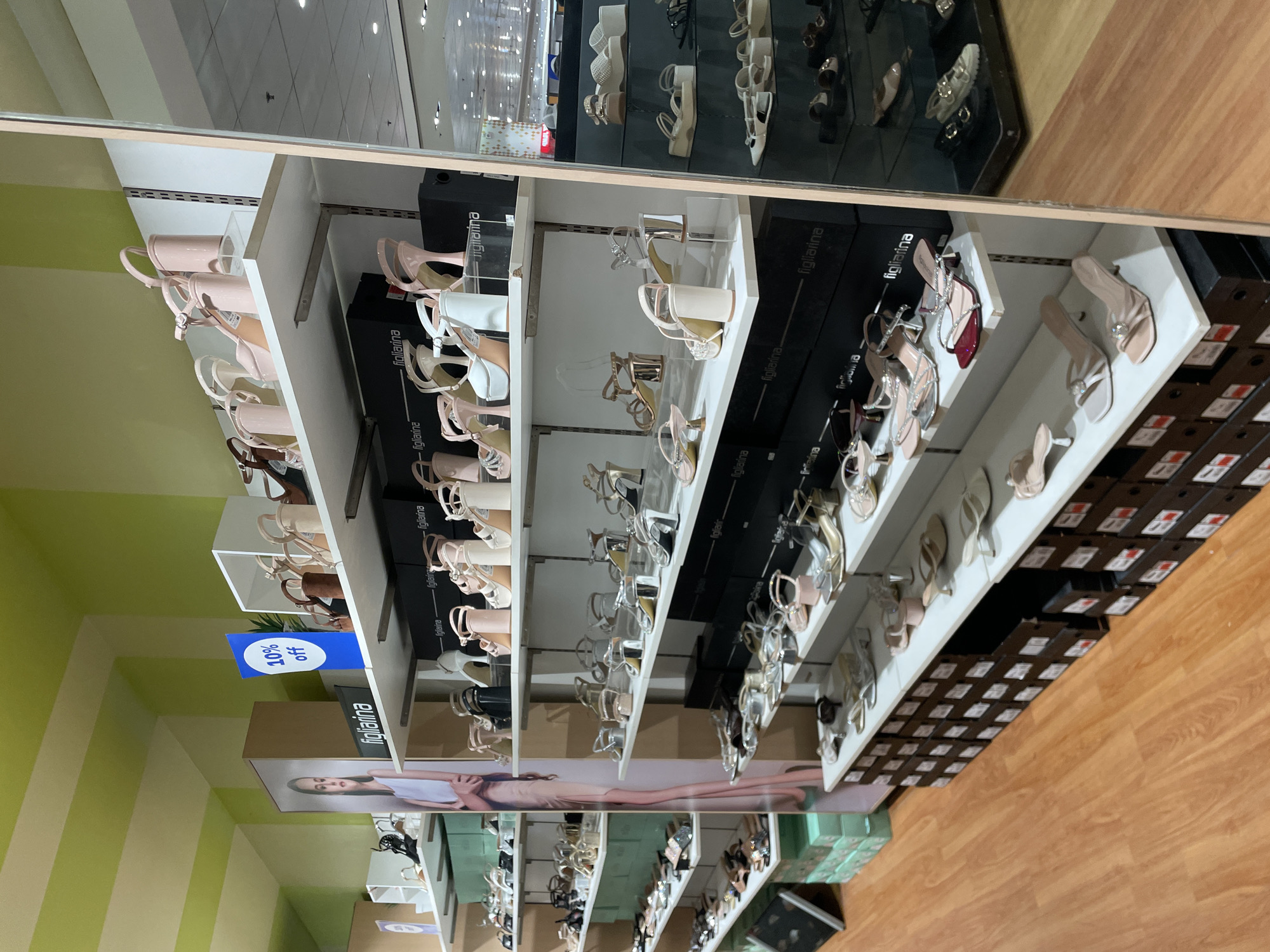 The image presents an interior view of a retail shoe store, featuring the "figliarina" brand, located in Davao City, Philippines. The scene is dominated by multiple white, multi-tiered display shelves showcasing a variety of women's footwear.
The shelves are laden primarily with high heels and sandals in diverse styles, including platform heels, block heels, and stilettos. Colors observed include various shades of nude, white, black, silver, gold, brown, and occasional red accents. Many shoes feature straps, buckles, or clear embellishments. The footwear is presented on clear acrylic risers or directly on the white shelves. Beneath the display shelves, numerous black shoe boxes, each marked with the "figliarina" brand name in white, are stacked, forming the lower structure of the displays. Additional green shoe boxes are visible on a shelf unit to the far left.
On the upper left, a large vertical advertising banner features a young woman with dark hair, shown from the chest up wearing a light top, and again from the mid-thigh down on a narrower section, modeling light-colored apparel. A blue circular sign with "10% off" in white text is prominently displayed on a shelf support in the upper center. A small potted green plant is partially visible behind this sign.
A tall, rectangular mirror on the far right edge of the frame reflects a portion of the adjacent retail space, revealing additional shoe displays featuring flatter sandals and loafers, along with overhead lighting and the store's ceiling. The store's ceiling and upper wall sections are painted with alternating horizontal stripes of light green and a lighter, off-white hue. The floor is constructed of polished light-brown wood planks. No individuals are actively present in the depicted scene, apart from the model on the banner.